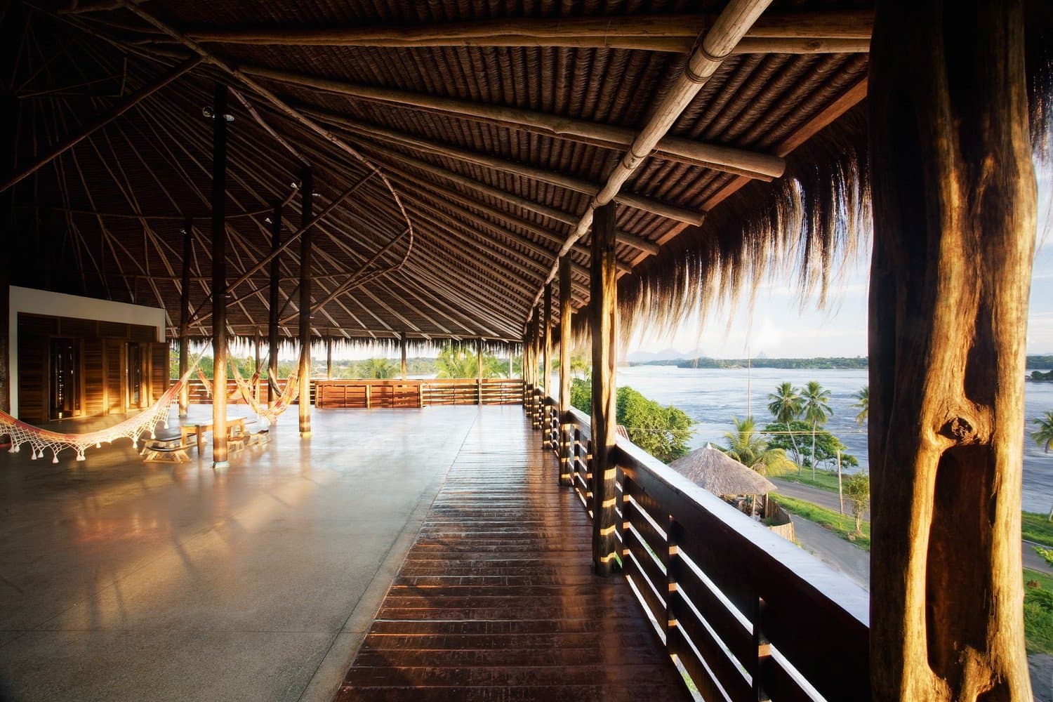 Hangmatten, warm licht en een rivier die je even doet vergeten wat er morgen op het programma staat. Photo: Daniel Ducci Open terras met rieten dak van een lodge aan de rivier tijdens de Pico da Neblina Expeditie, met hangmatten tussen houten palen en een weids uitzicht over een brede rivier met palmen in het gouden-uurlicht