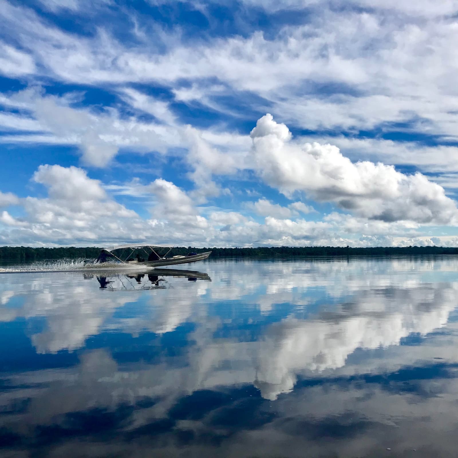 Glad water, hoge lucht, en het gevoel dat de rivier nooit ophoudt. Een gemotoriseerde kano scheurt over een spiegelgladde zwartwater rivier tijdens de Pico da Neblina Expeditie, met een dicht woud aan de horizon en grote stapelwolken die perfect weerkaatsen op het wateroppervlak