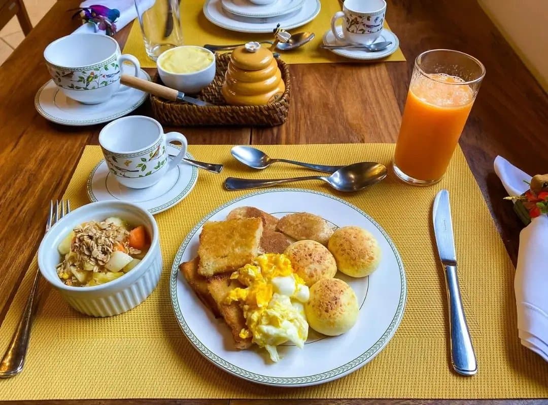 Du pão de queijo dès le matin. Le reste de la journée n'a plus qu'à suivre. Un petit-déjeuner généreux dans un lodge du Pantanal Sud avec œufs brouillés, pain grillé, pão de queijo, jus d'orange frais, granola aux fruits, miel et tasses à café à motifs floraux sur un set de table jaune