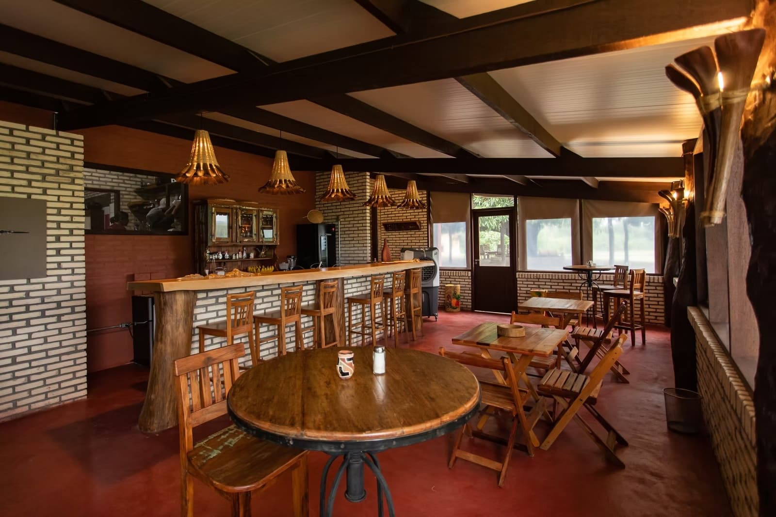 Bois chaud, abat-jours tressés, et un verre frais qui attend au retour du terrain. Salle à manger et bar rustiques d'un lodge de safari dans le sud du Pantanal, avec des tables rondes en bois, des tabourets hauts le long d'un comptoir en brique, des suspensions artisanales et de grandes fenêtres donnant sur la végétation