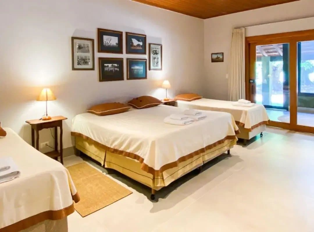 Draps propres, lumière douce et verdure à portée de main. Profitez-en, les fourmiliers géants lèvent tôt. Chambre triple dans un lodge de safari au Pantanal sud, avec trois lits aux draps crème et caramel, des lampes de chevet chaleureuses, des photographies en noir et blanc encadrées au mur et des portes coulissantes en verre donnant sur le jardin