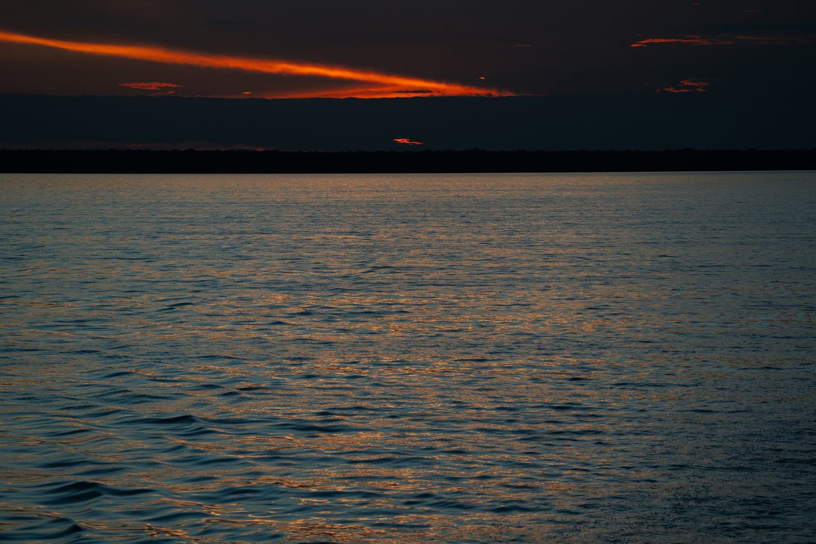 Het laatste licht van de dag, en de rivier houdt het allemaal vast. Dieprode en oranje wolkenstrepen gloeien boven een brede, donkere rivier in de schemering tijdens de Tucano Amazon-riviercruise, met een beboste horizon als silhouet onder een bijna zwarte lucht