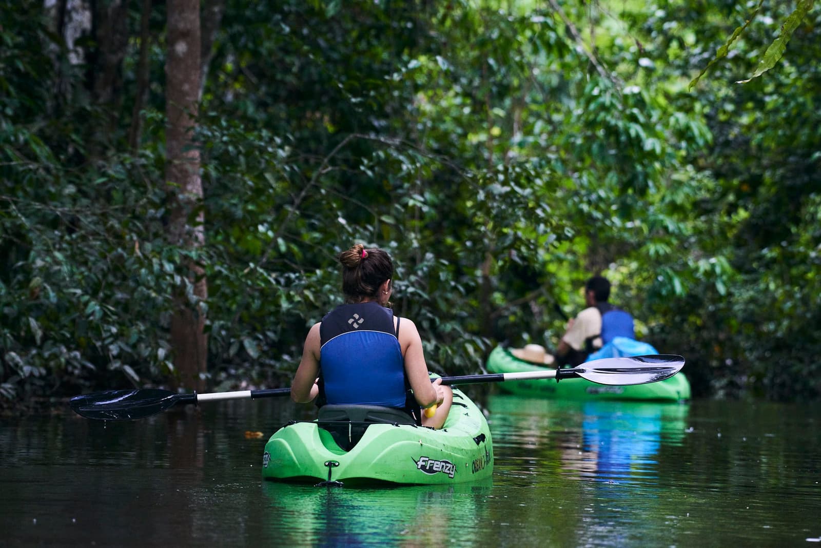 Donker water, gesloten bladerdak, nergens een verplichting. Gewoon peddelen. Photo: JUKAPAZ Twee kajakkers peddelen in groene kajaks door een smal, donker waterkanaal omgeven door dicht regenwoud tijdens een Tucano Amazon River Cruise igapó-uitstap