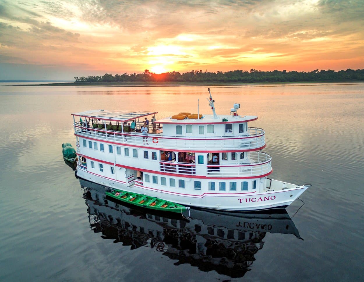Vijf nachten je thuis op het water, met uitzicht inbegrepen. Het M/Y Tucano, een wit driedeks riviercruiseschip, voor anker op een spiegelgladde donkerwater rivier bij zonsondergang, met een groene houten kano ernaast en passagiers op het bovendek, bossilhouet tegen een oranje lucht