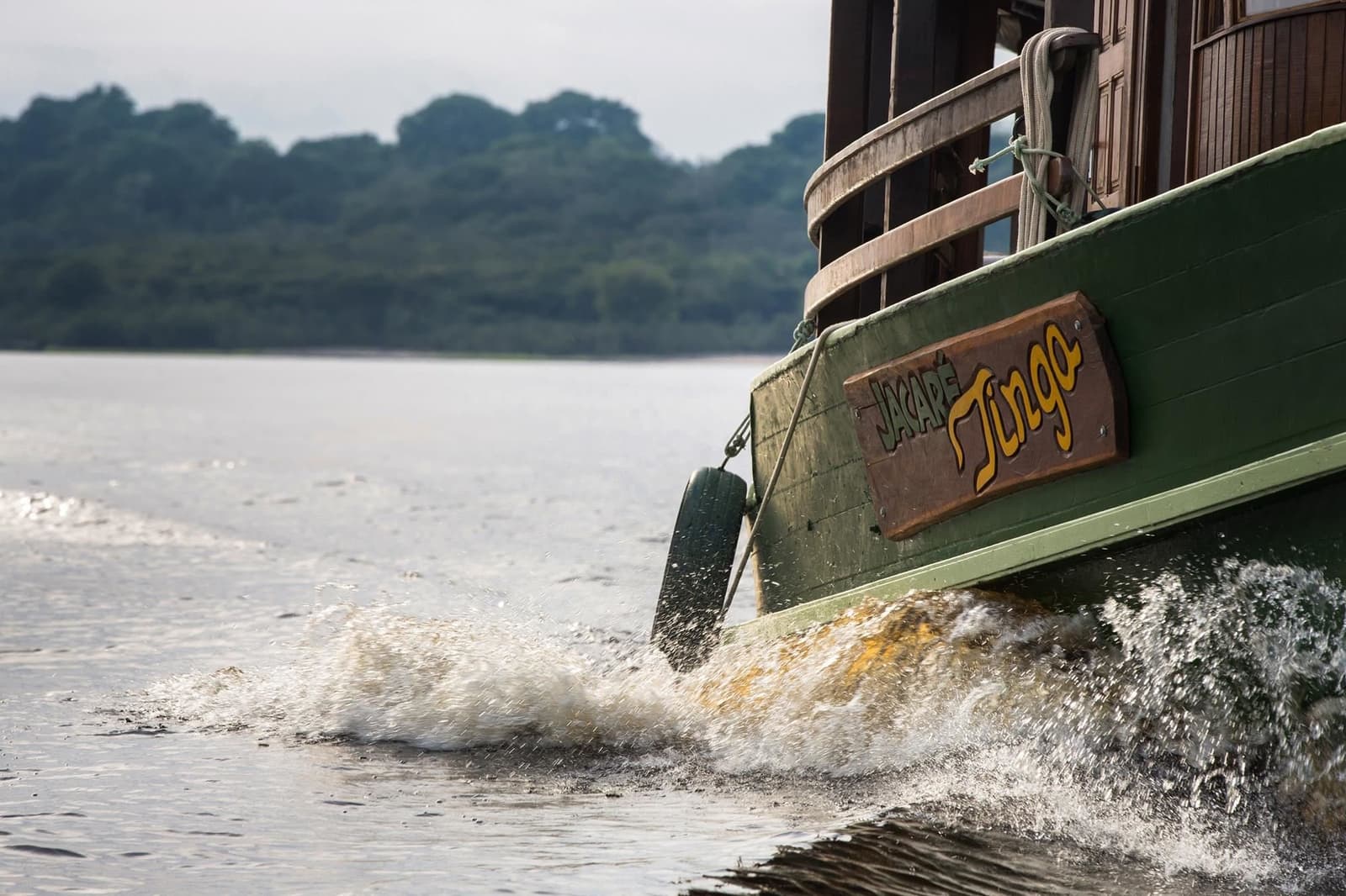 O Jacaré Tingo conhece cada curva desse rio de cor. A proa do barco fluvial Jacaré-Açu cortando as águas escuras do rio Amazônico, levantando espuma branca, com floresta densa na margem oposta