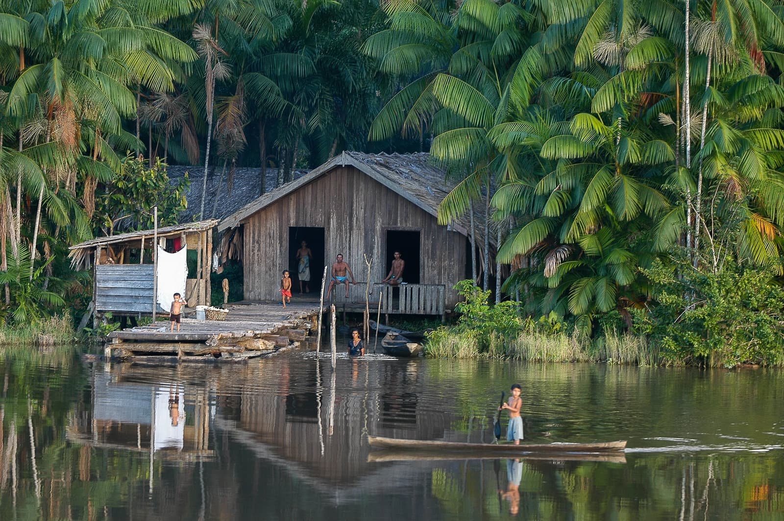 O rio dita o ritmo, e a vida segue sem pressa. Uma casa de madeira sobre palafitas à beira de um rio na Amazônia, cercada por densa floresta de palmeiras, com uma família na varanda e no trapiche, uma pessoa caminhando na água rasa e uma criança em pé numa canoa no rio calmo — um recorte da vida ribeirinha em um cruzeiro pela Amazônia no Brasil