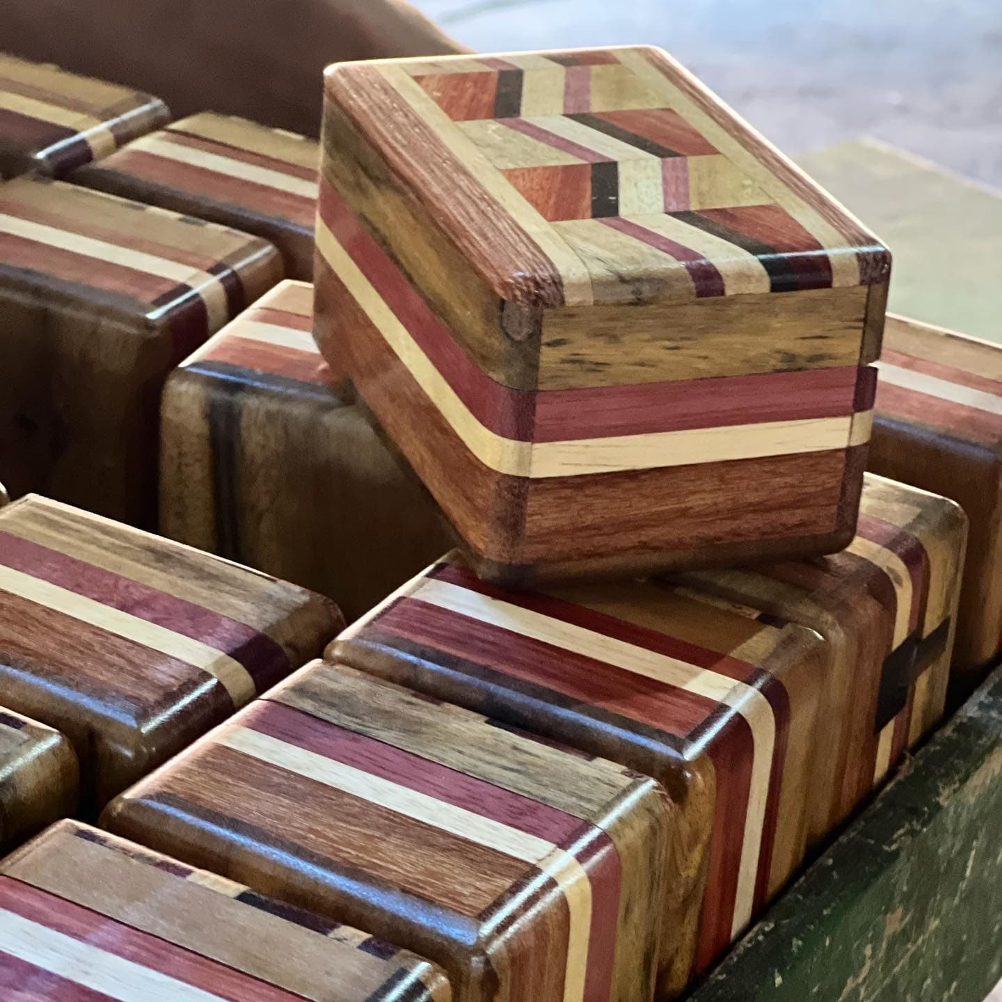 Pas une boîte n'est identique à sa voisine, et c'est tout le charme de l'affaire. Collection de petites boîtes en bois artisanales ornées de motifs rayés incrustés dans des tons de rouge chaud, crème et brun foncé, fabriquées à partir de plusieurs essences de bois et exposées dans un plateau lors d'une halte en atelier artisanal
