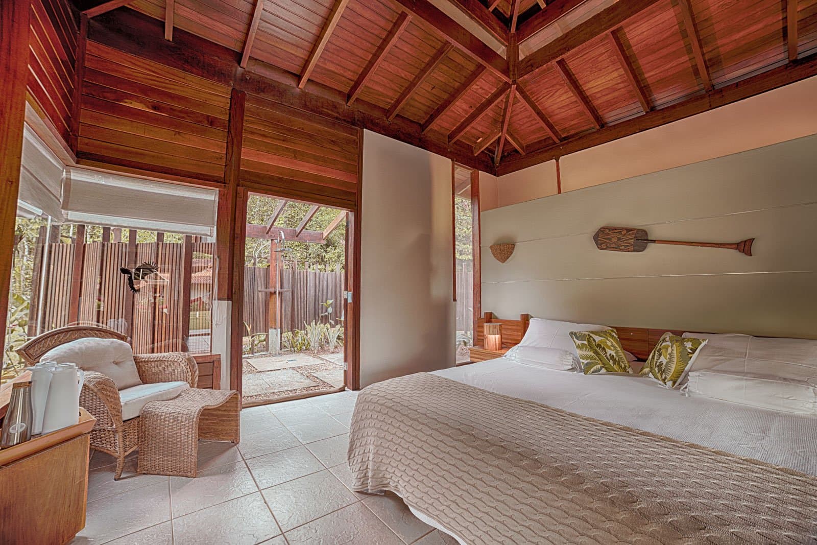 Paddle on the wall, garden out the door, birds queuing up at first light. Photo: Samuel Melim Spacious bungalow bedroom with a vaulted hardwood ceiling, a double bed dressed in tropical-print cushions, wicker armchairs, and open sliding doors leading to a private outdoor shower garden, at a lodge suited for birdwatching in the Amazon rainforest