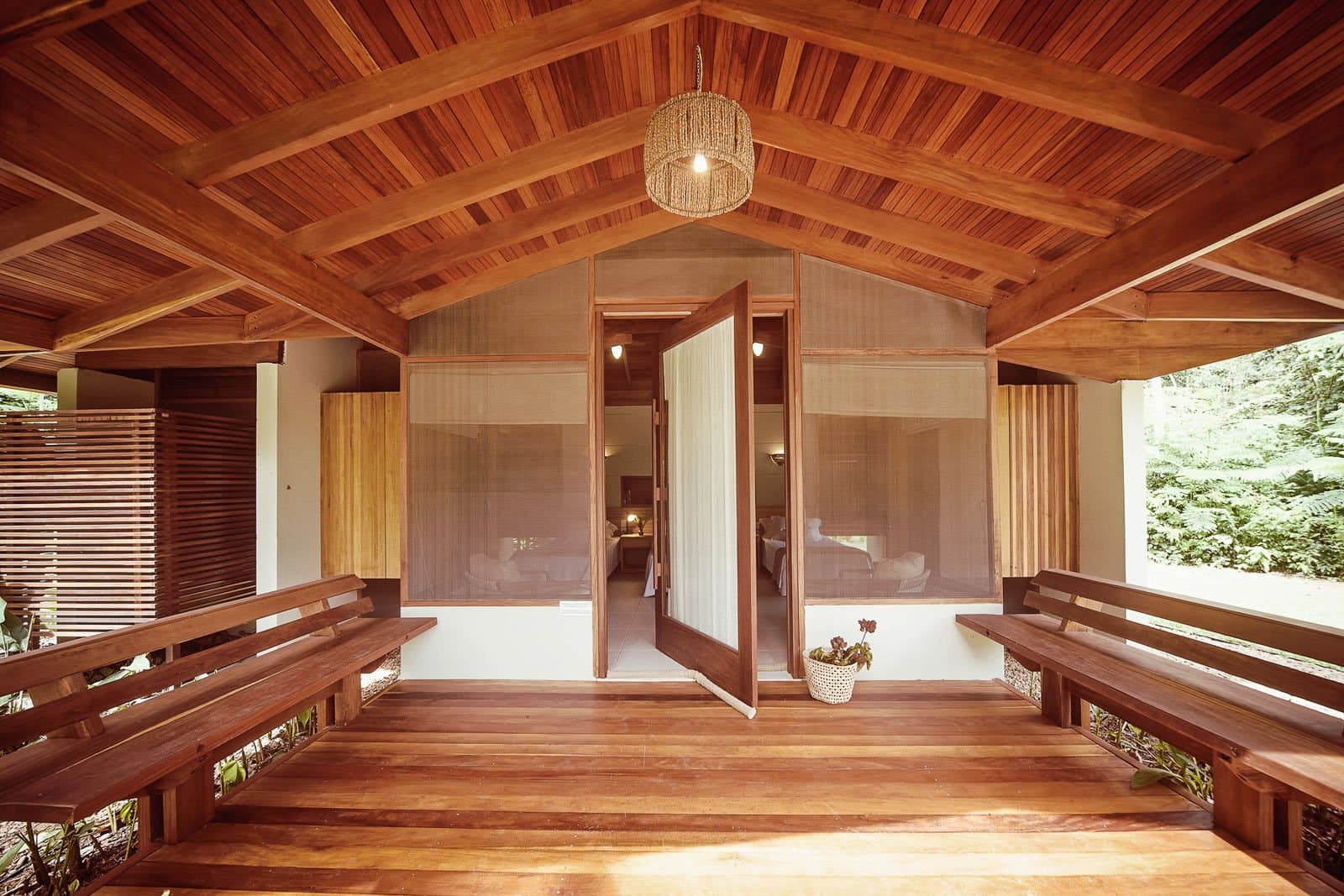 Wood this good makes it hard to leave for the 5 AM trail. Photo: | SamuelMelim Photography Warm timber veranda of a jungle bungalow at a lodge for birdwatching in the Amazon rainforest, with two wooden bench seats, a slatted ceiling, a woven pendant light, and an open door revealing a twin bedroom beyond dense green forest