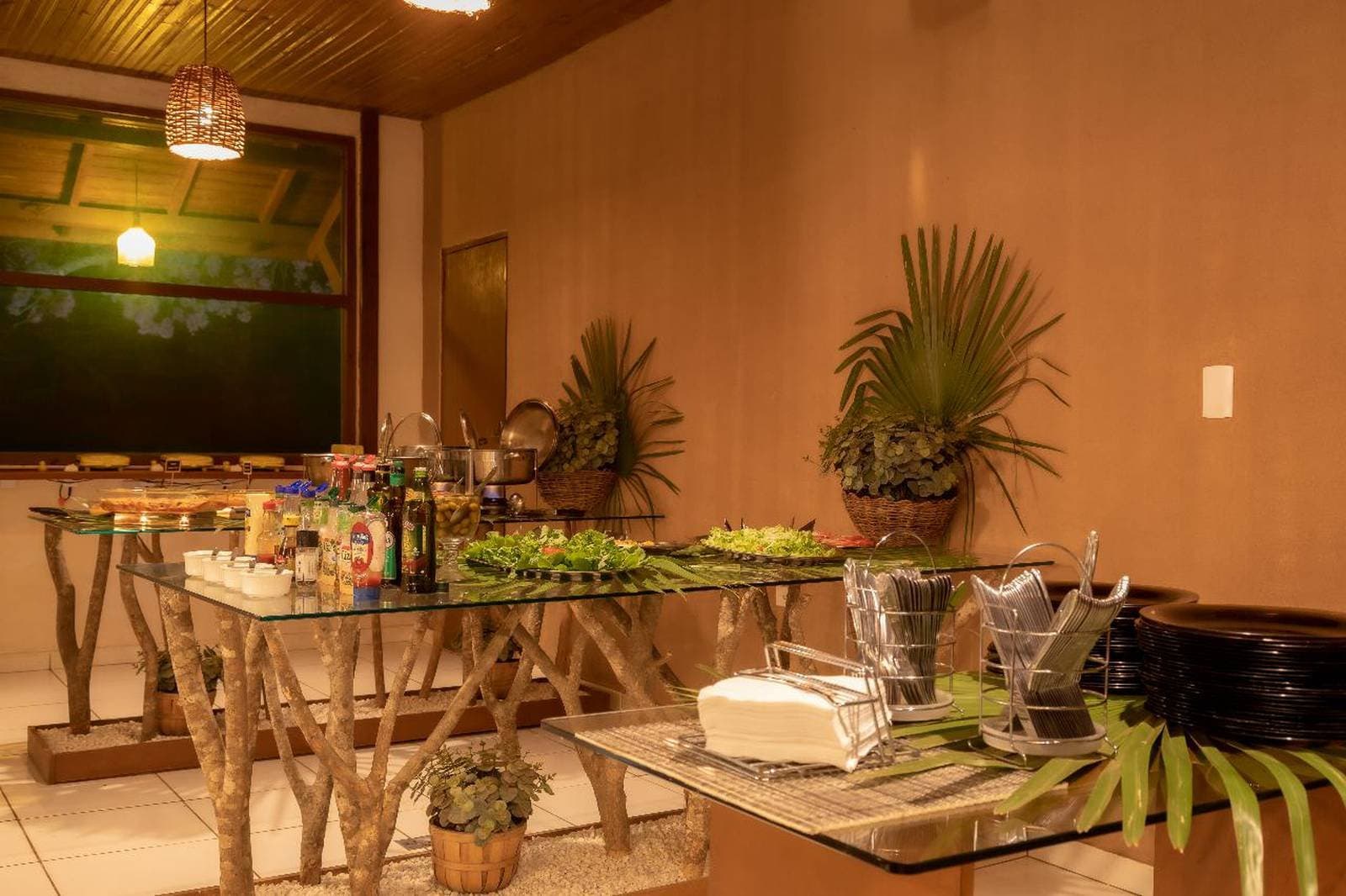 Nach einem langen Tag draußen schmeckt das Abendessen einfach besser. Innen-Buffet in einer Pantanal-Budget-Lodge mit Glastischen auf rustikalen Astbeinen, beladen mit Salaten, Gewürzen, warmen Speisen und tropischen Pflanzen unter warmem Hängeleuchten