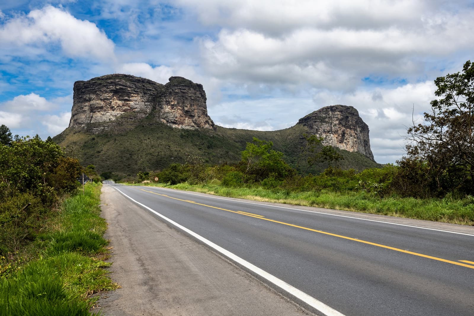 La route est vide, les falaises sont immenses, et le silence fait le reste. Une route à deux voies déserte mène tout droit vers deux mesas de grès aux sommets plats se dressant au-dessus d'une végétation verte lors d'un circuit Chapada Diamantina