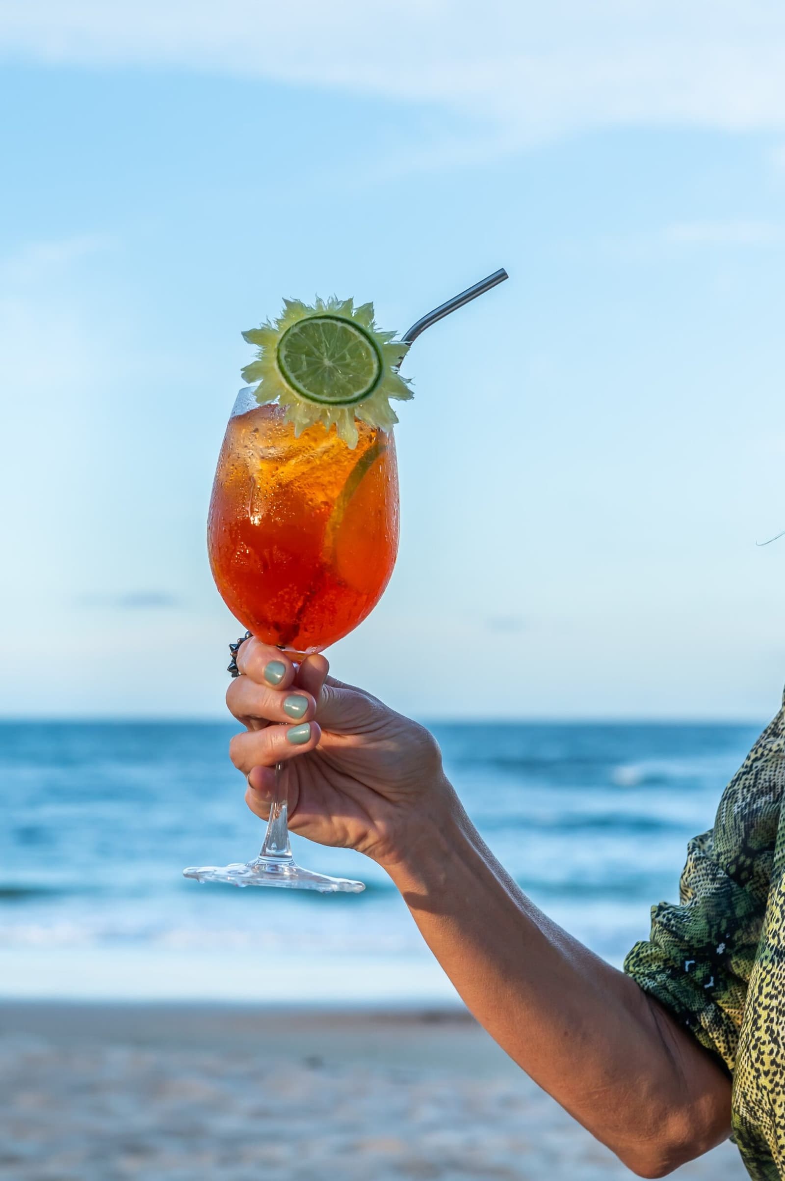 Merecido. Drinque na mão, mar na frente, roteiro cumprido. Uma mão segura uma taça grande de drinque vermelho gelado, enfeitado com rodela de limão e ervas, com o mar ao fundo na costa da Bahia