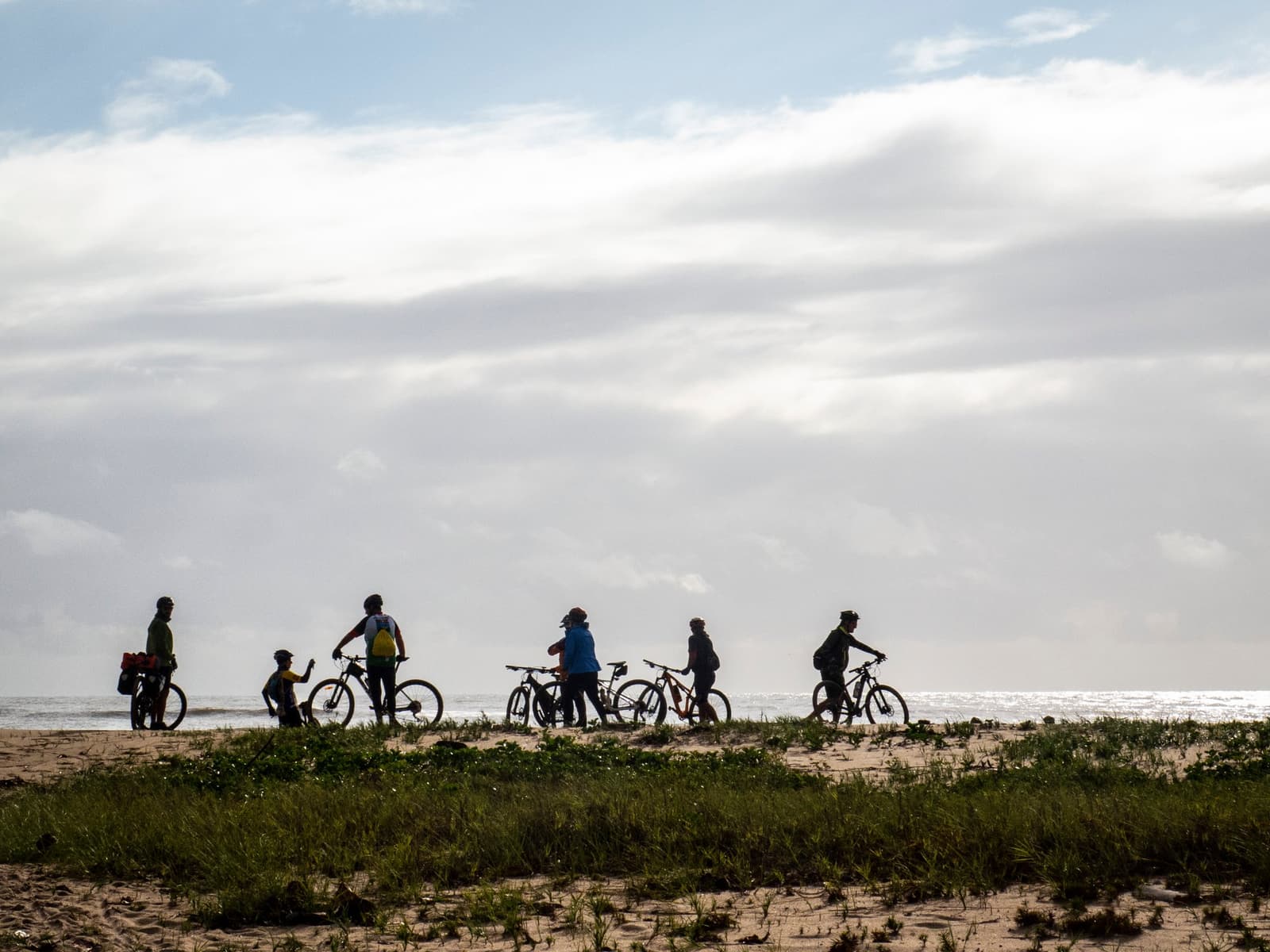 Seis ciclistas, um horizonte, e a rota inteira esperando por eles. Grupo de ciclistas com bicicletas de montanha parados sobre uma duna costeira coberta de vegetação rasteira, em silhueta contra o céu nublado na rota de cicloturismo pela Bahia