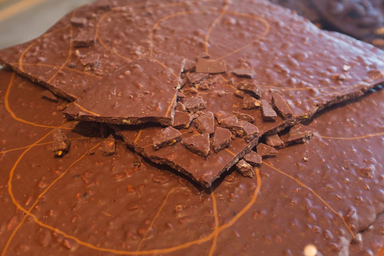 Quebra, prova, repete. O chocolate da Fazenda Vila Rosa dispensa qualquer cerimônia. Pedaços de chocolate artesanal com inclusões de castanha espalhados sobre uma placa fina de chocolate em superfície marmorizada, durante degustação em fazenda de cacau na rota de ciclismo pela Bahia