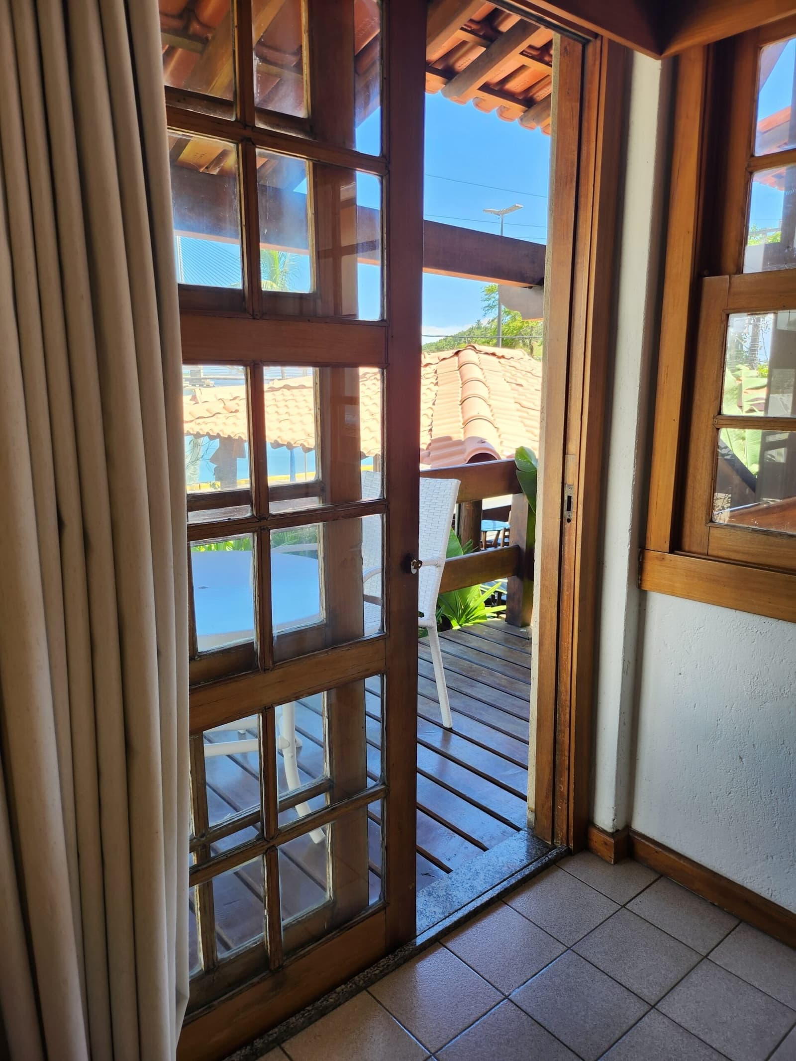 Porta aberta, deck à espera. Às vezes é simples assim. Vista através de uma porta de madeira com vidro aberta para um deck de madeira ensolarado com cadeira branca, telhas de barro e vegetação tropical em uma pousada