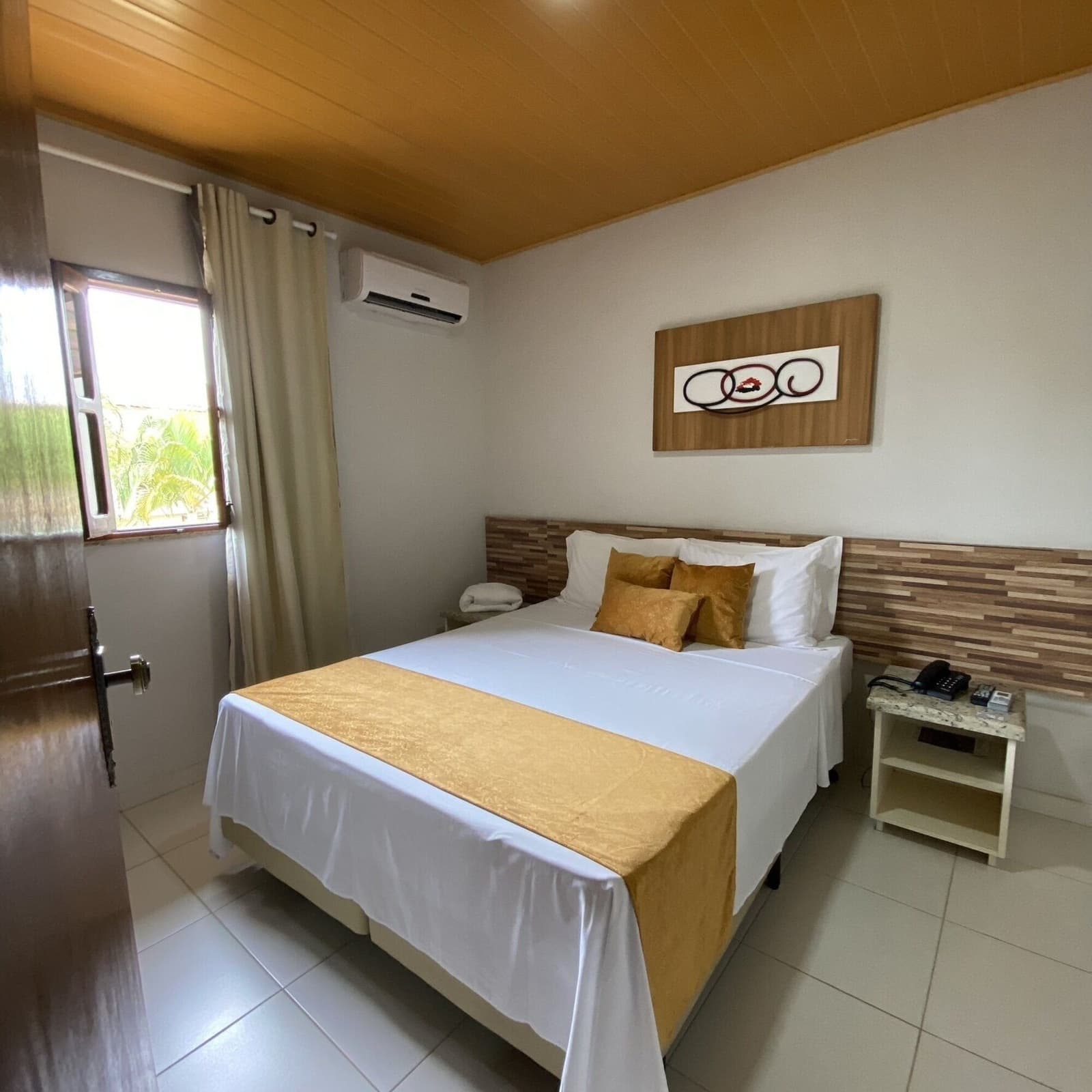 Um quarto simples e bem cuidado, que cai muito bem após o dia pedalando. Cama de casal arrumada com almofadas e caminho de cama em tom dourado em quarto de hotel climatizado com forro de madeira no teto e janela com vista para vegetação tropical, acomodação em roteiro de cicloturismo na Bahia