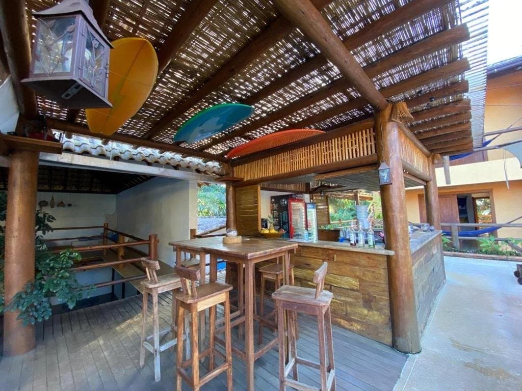 Banquetas livres, bebidas geladas, caiaques no teto, alguém acertou nas prioridades. Bar ao ar livre com estrutura em madeira rústica, teto de bambu, banquetas altas e caiaques coloridos pendurados no teto em uma pousada na rota de ciclismo na Bahia