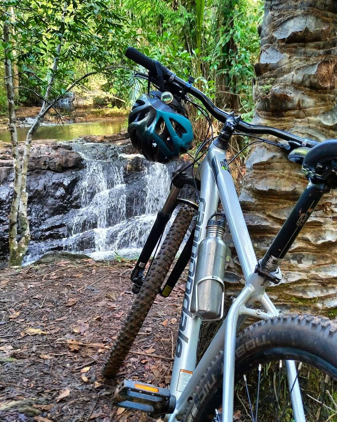 Tem parada que a trilha organiza por conta própria. Uma mountain bike com capacete azul-petróleo apoiada em uma rocha de arenito ao lado de uma pequena cachoeira em cascata em uma trilha de ciclismo na Bahia, em meio à Mata Atlântica