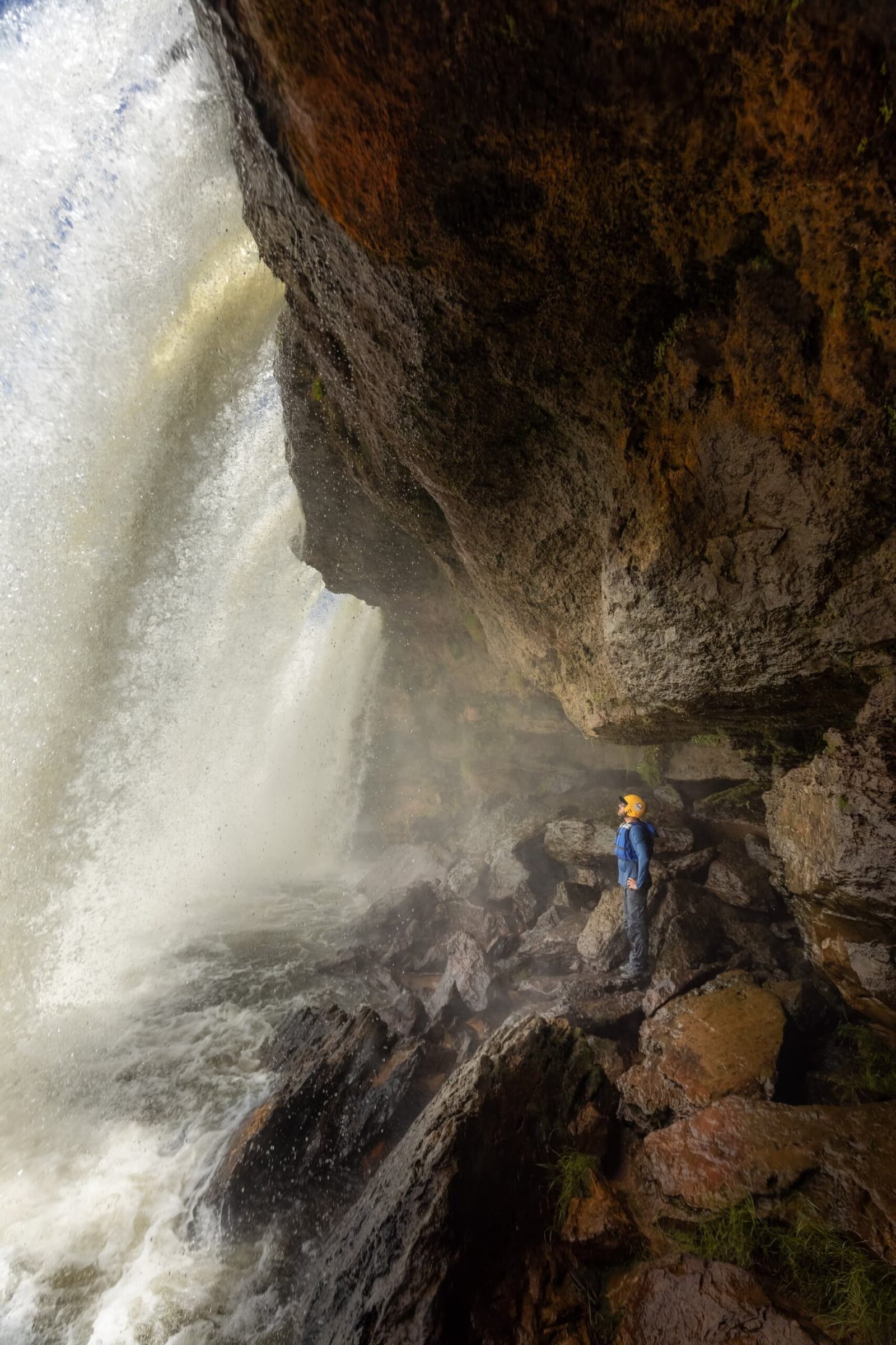 O trovão da queda chega primeiro, bem antes de você a ver. Photo: Alexandre Socci Um aventureiro de capacete e jaqueta azul parado num patamar rochoso sob uma cachoeira em queda livre durante uma aventura no Jalapão, envolto em névoa e paredes de rocha cor de ferrugem