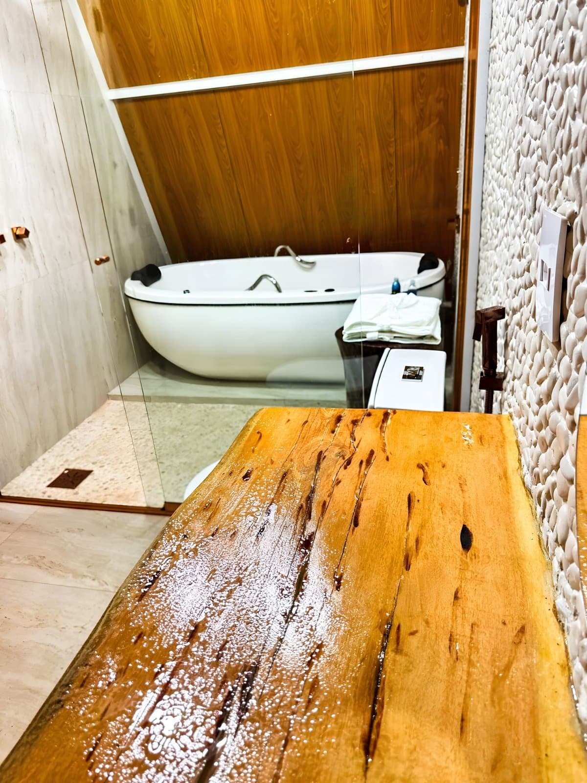 Tired muscles, meet your match. The lodge bathroom has other plans for you tonight. A spa-style bathroom with a freestanding white soaking tub set against warm wood-panelled walls, a textured pebble stone accent wall, and a wet live-edge wooden bench in the foreground, at a lodge on a Jalapão trekking tour