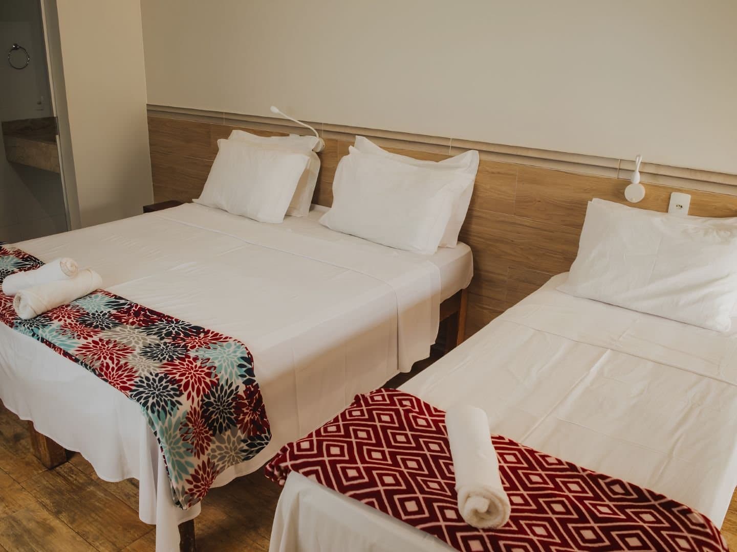 Three beds, fresh towels, and zero complaints after a day on dirt roads. A clean triple guestroom on a Jalapão trip, with two double beds and a single bed dressed in white linen, colorful bed runners, rolled towels, a wooden headboard panel, and a wood-plank floor.