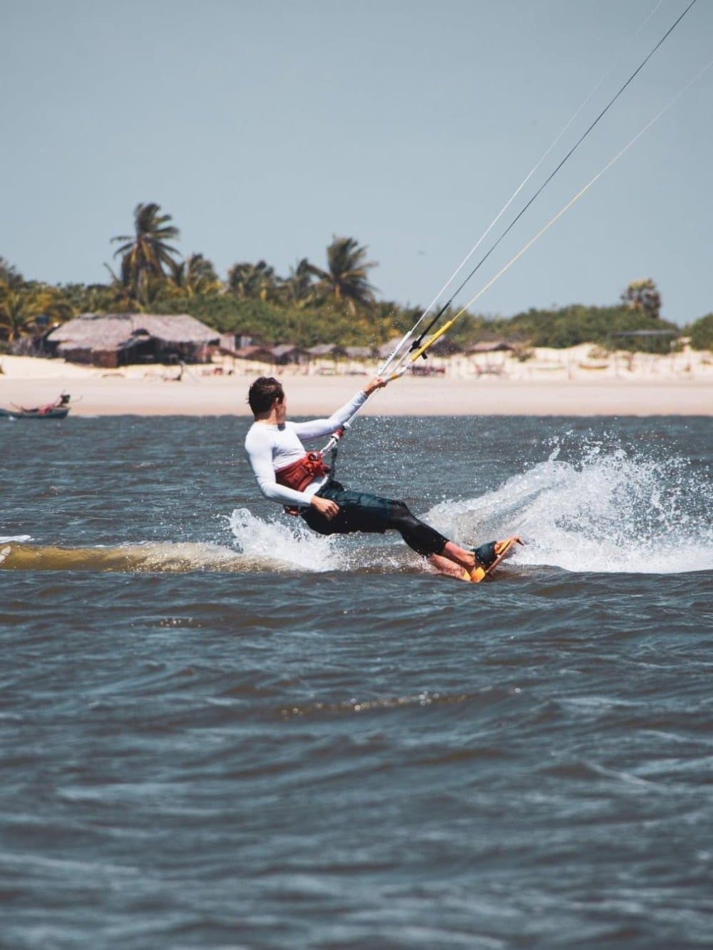 O vento dos Lençóis não pede licença para te levar junto. Um kitesurfista desliza sobre águas agitadas próximo a uma praia de areia branca ladeada de coqueiros e casas com telhado de palha