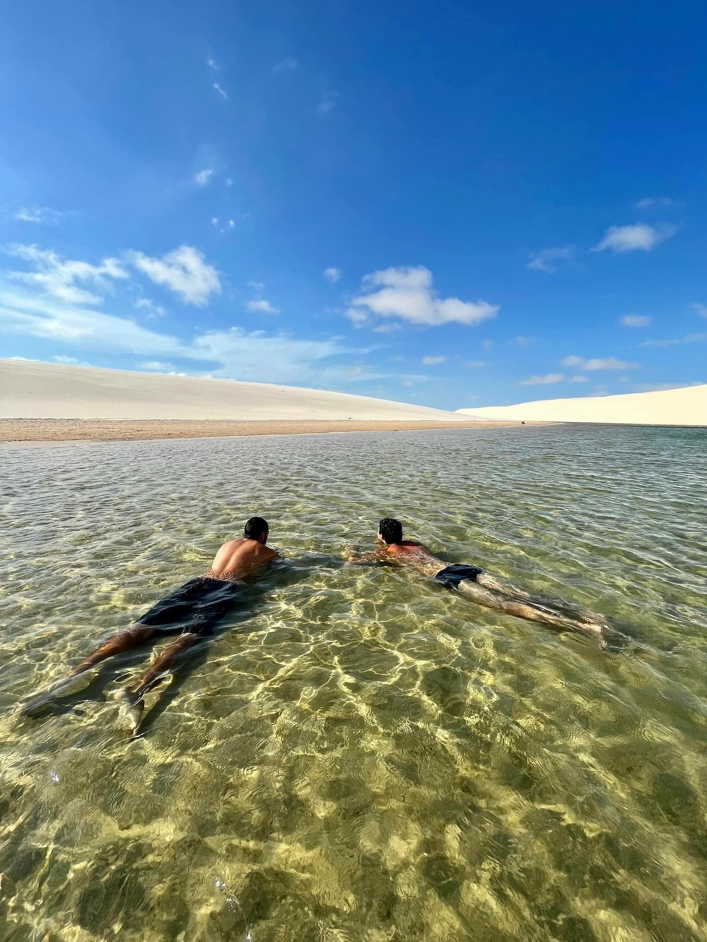 A água tão transparente que parece que você está voando sobre a areia. Dois banhistas flutuam de bruços em uma lagoa rasa e cristalina nos Lençóis Maranhenses, com dunas de areia branca se estendendo até o horizonte sob um céu azul intenso