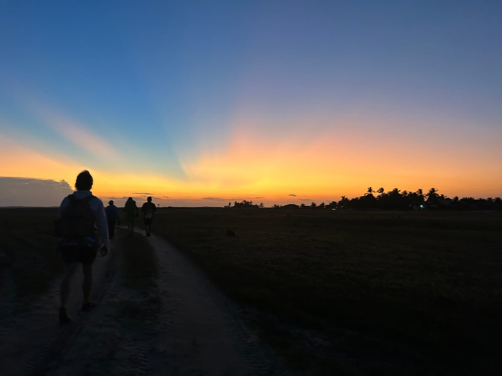 The last light of the day belongs to whoever keeps walking. Silhouettes of hikers walking along a sandy trail toward a vivid orange and blue sunset sky on a Lençóis Maranhenses hike, with palm trees on the horizon