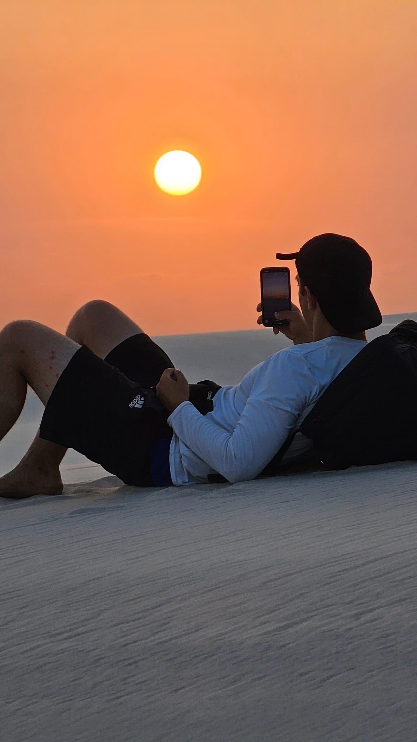 Certains couchers de soleil méritent qu'on s'allonge dans le sable pour les savourer. Un voyageur allongé sur une dune de sable blanc à Lençóis Maranhenses photographiant un grand soleil orange à l'horizon avec son smartphone