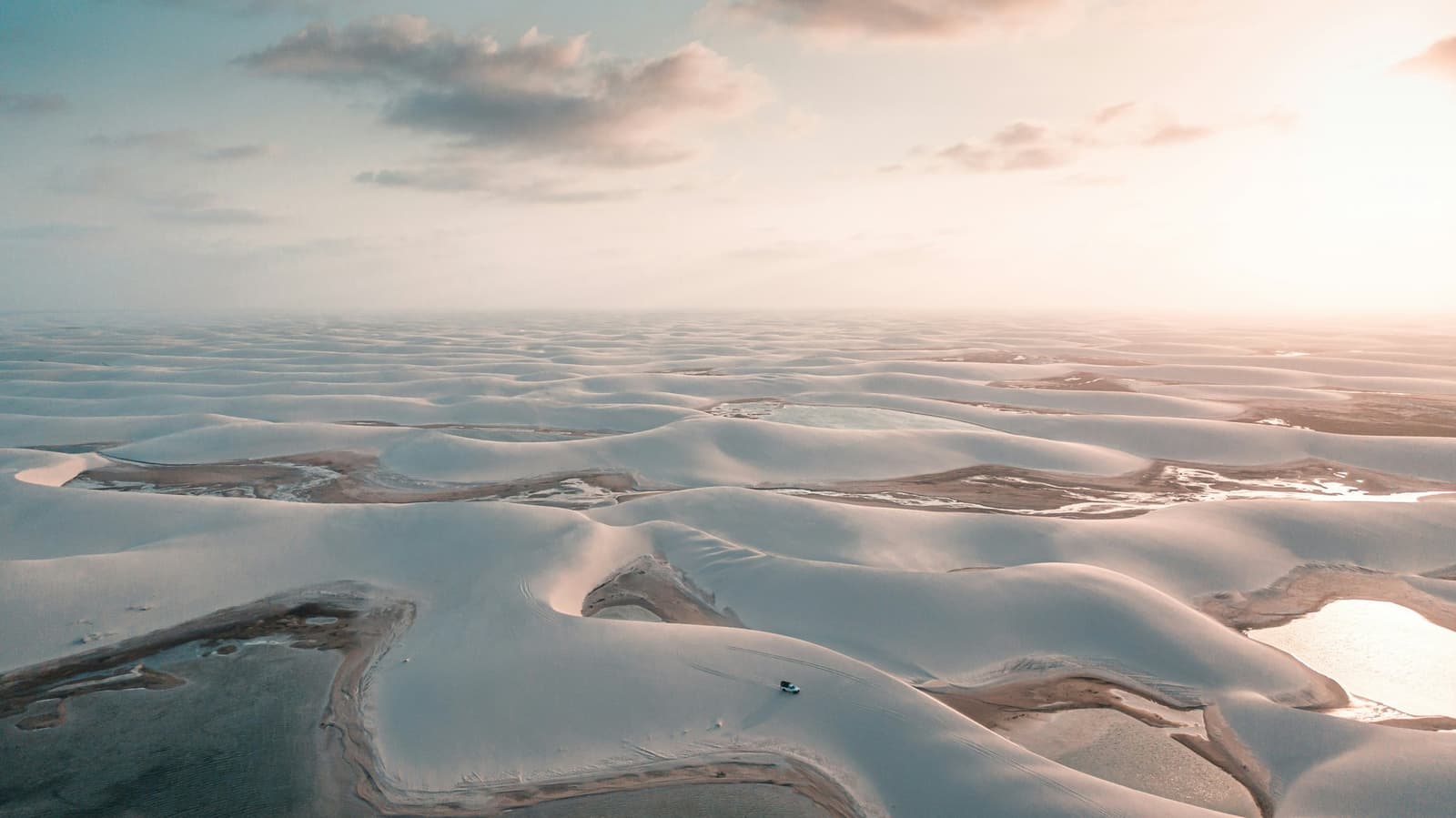 Dunas sem fim, algumas lagoas e um carro minúsculo para lembrar a escala de tudo isso. Vista aérea do trek nos Lençóis Maranhenses saindo de Atins, com um único 4x4 atravessando uma imensidão de dunas brancas salpicadas de lagoas rasas sob um céu dourado