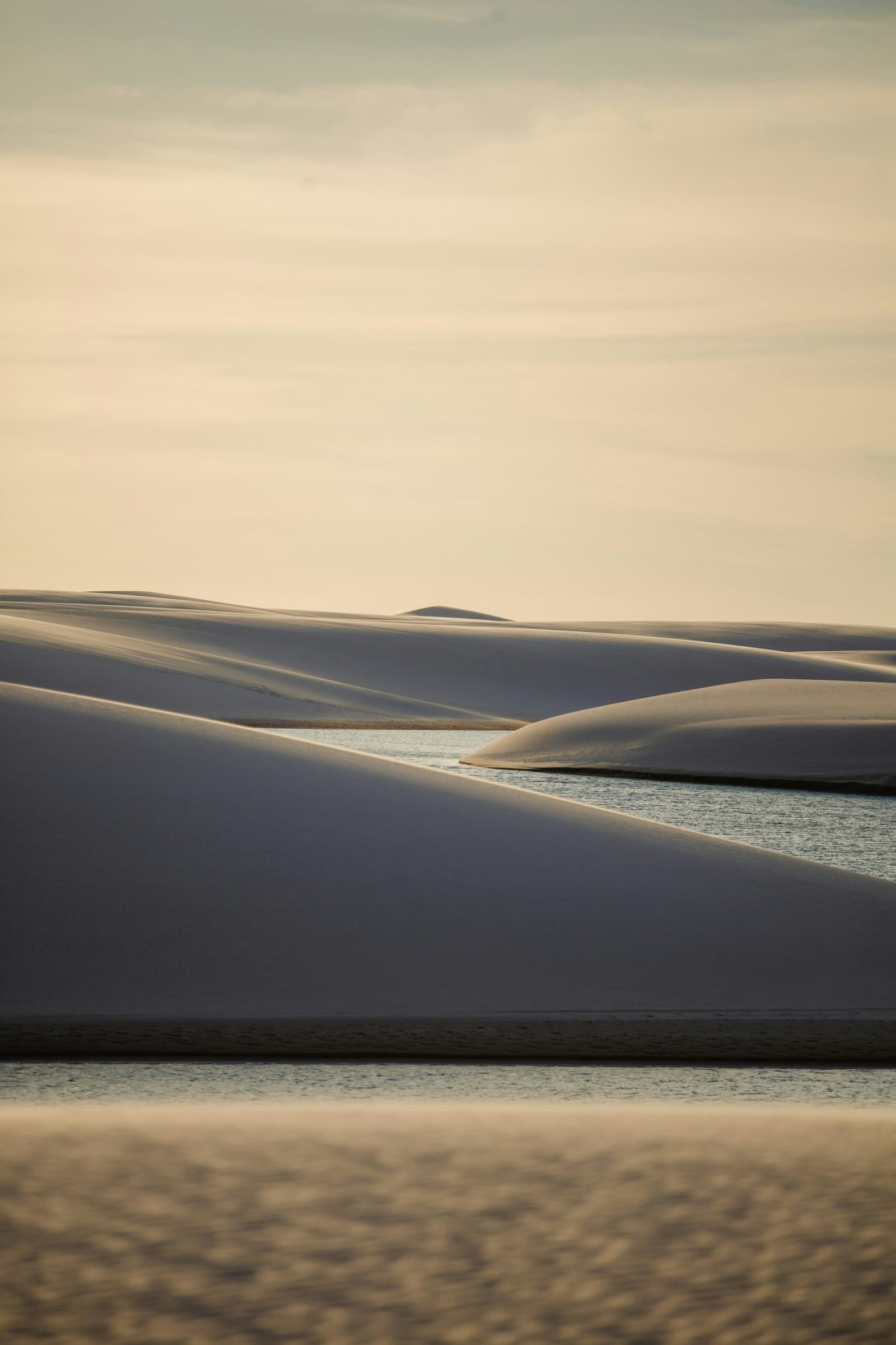 Areia que parece esculpida, água que surge do nada. Dunas de areia branca do trek pelos Lençóis Maranhenses projetando sombras azuladas sobre uma estreita lagoa de água doce sob a luz dourada do entardecer, com céu claro e velado ao fundo