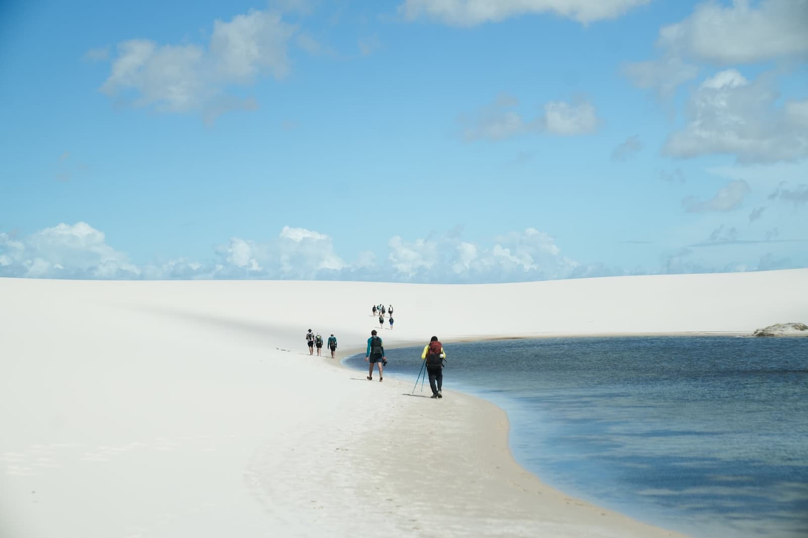 Le sentier longe la lagune, et personne ne se plaint. Un groupe de randonneurs équipés de sacs à dos marche en file indienne sur une bande de sable blanc entre une lagune bleue et de grandes dunes lors d'un trek dans les Lençóis Maranhenses sous un ciel partiellement nuageux
