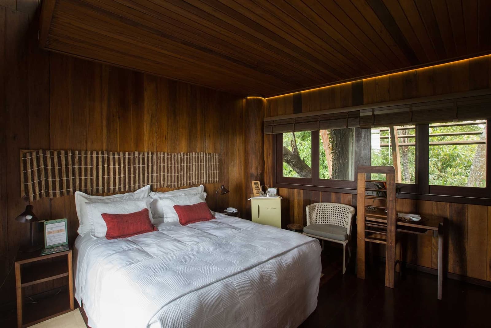 Du bois chaud, la forêt à portée de regard, et un lit qui appelle au repos. Chambre du lodge Mirante do Gavião avec murs en bois sombre, literie blanche immaculée, coussins rouges et fenêtres donnant sur la forêt amazonienne