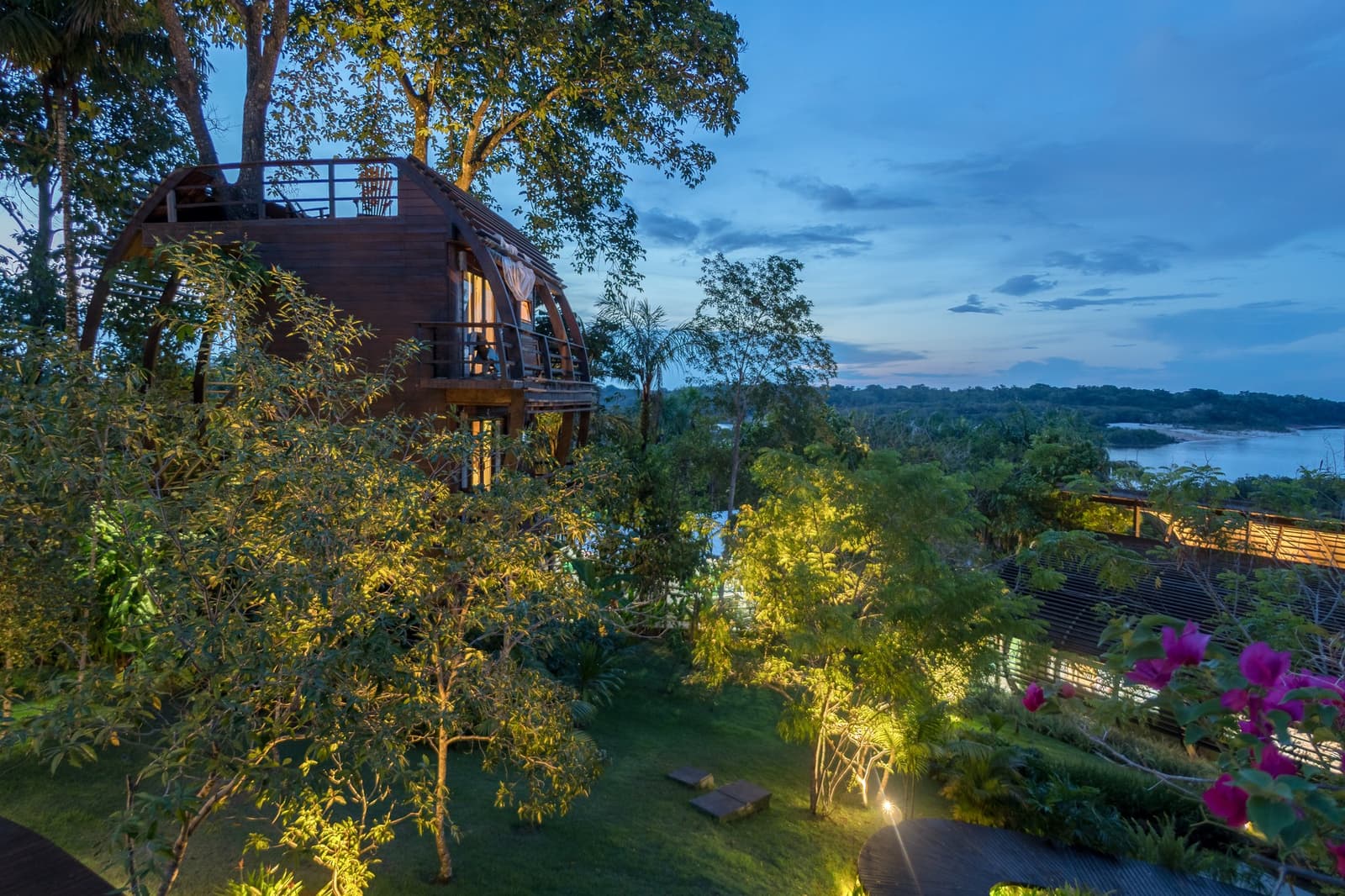 Une chambre perchée dans la canopée, le fleuve en fond de tableau. Une suite en bois en forme de dôme au Mirante do Gavião brille d'une lumière chaleureuse au crépuscule, entourée d'un jardin tropical luxuriant avec vue sur une large courbe de rivière sous un ciel bleu nuit