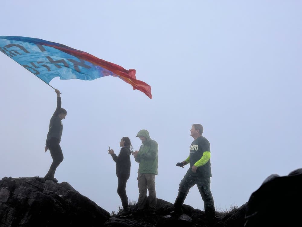 De vlag omhoog, de wolken dichterbij, en niemand die er iets op tegen heeft. Vier wandelaars als silhouet in dichte mist op een rotsachtige kam tijdens een Pico da Neblina Expeditie, één persoon heft een grote kleurrijke vlag die wappert in de wind