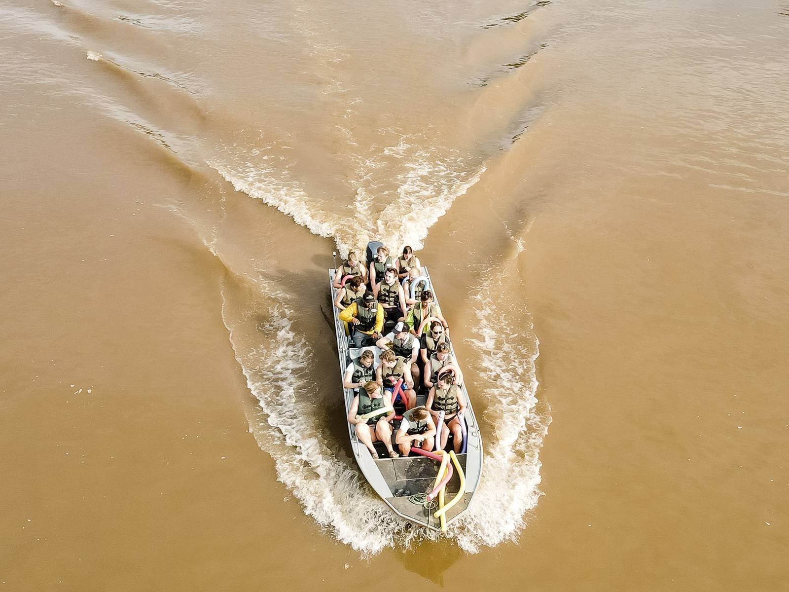 Bateau plein, rivière large, lodge encore loin. Ça commence bien. Vue aérienne d'un hors-bord bondé filant sur un fleuve aux eaux boueuses lors d'un safari dans le sud du Pantanal, passagers en gilets de sauvetage, large sillage en V derrière l'embarcation