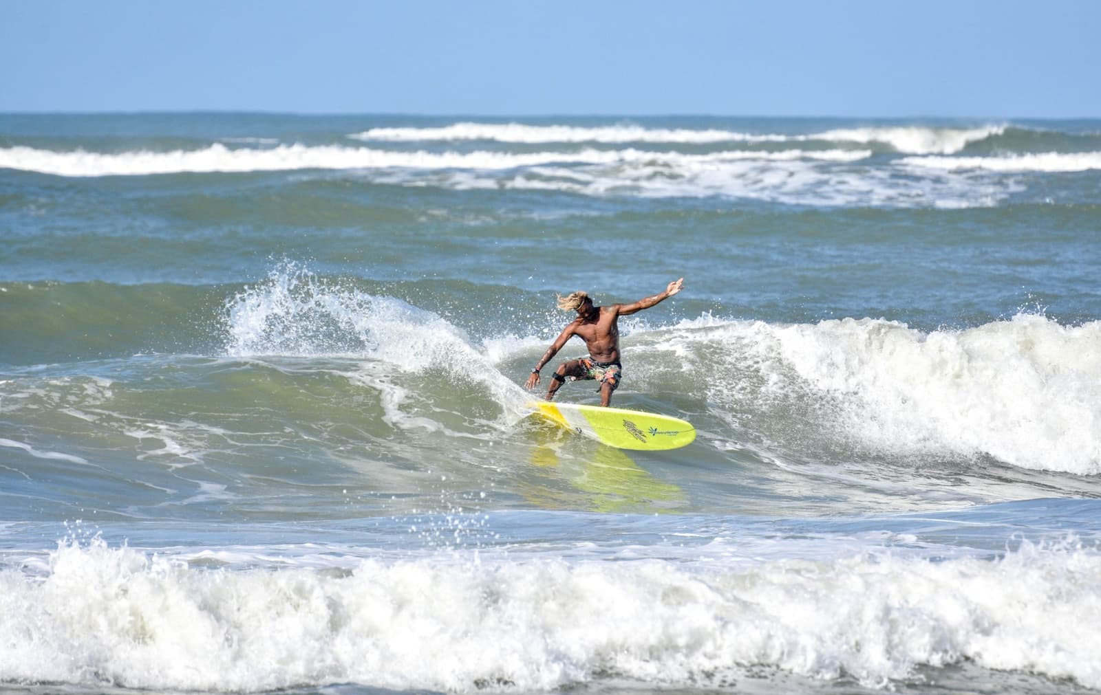 Armen wijd, haren los, volledig opgegaan in de golf. Dat is het doel. Photo: Picasa Een surfer met lang blond haar rijdt op een geel longboard door een brekende golf tijdens surflessen in Itacaré, armen gespreid voor balans onder een heldere blauwe lucht.