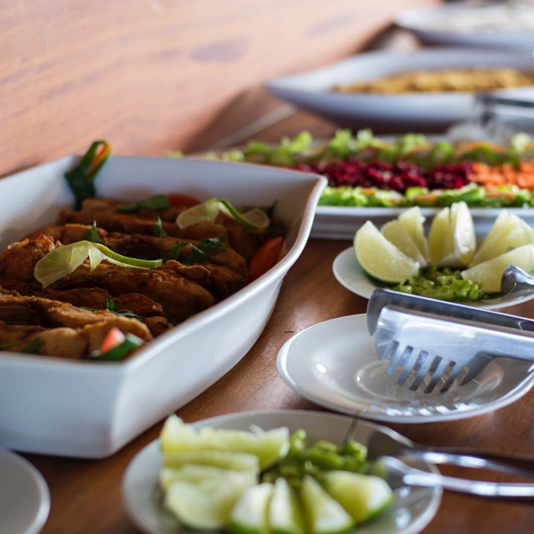 Comida caseira, rio à janela. Não falta nada. Um buffet servido a bordo de um cruzeiro pelo Amazonas no Brasil, com uma travessa de peixe frito dourado com rodelas de limão e ervas, pratos coloridos de salada com beterraba, cenoura e folhas verdes, e pratinhos de pepino e limão sobre uma mesa de madeira
