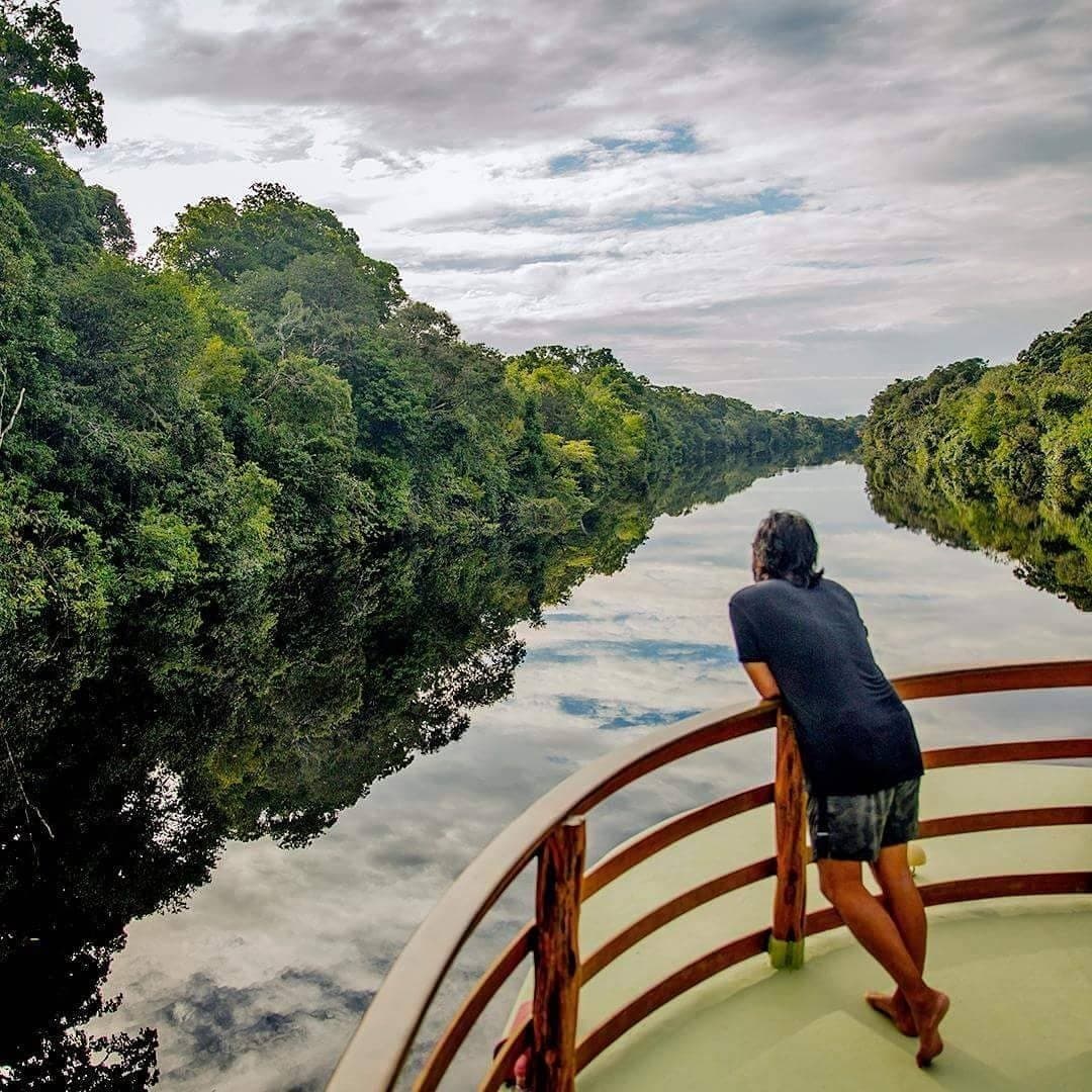 Devant vous, juste la forêt, l'eau noire et le silence du fleuve. Un passager s'appuie sur le bastingage en bois de la proue d'un bateau fluvial lors d'une croisière sur le fleuve Amazone au Brésil, contemplant un canal d'eau noire parfaitement lisse bordé de forêt tropicale dense