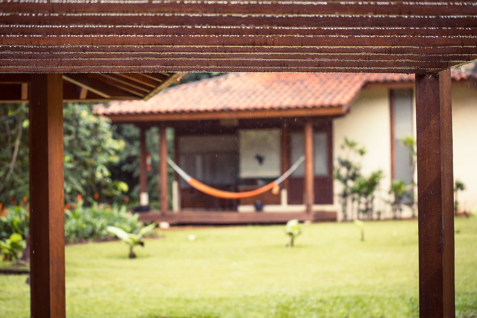 La pluie sur les tuiles, le hamac qui attend. Certains après-midis s'imposent d'eux-mêmes. Photo: | SamuelMelim Photography Une pergola en bois au premier plan cadre la vue sur un bungalow de lodge avec un toit en tuiles de terre cuite, un hamac orange sur la véranda et une pelouse verte sous la pluie — un refuge entre deux sorties d'observation des oiseaux en Amazonie