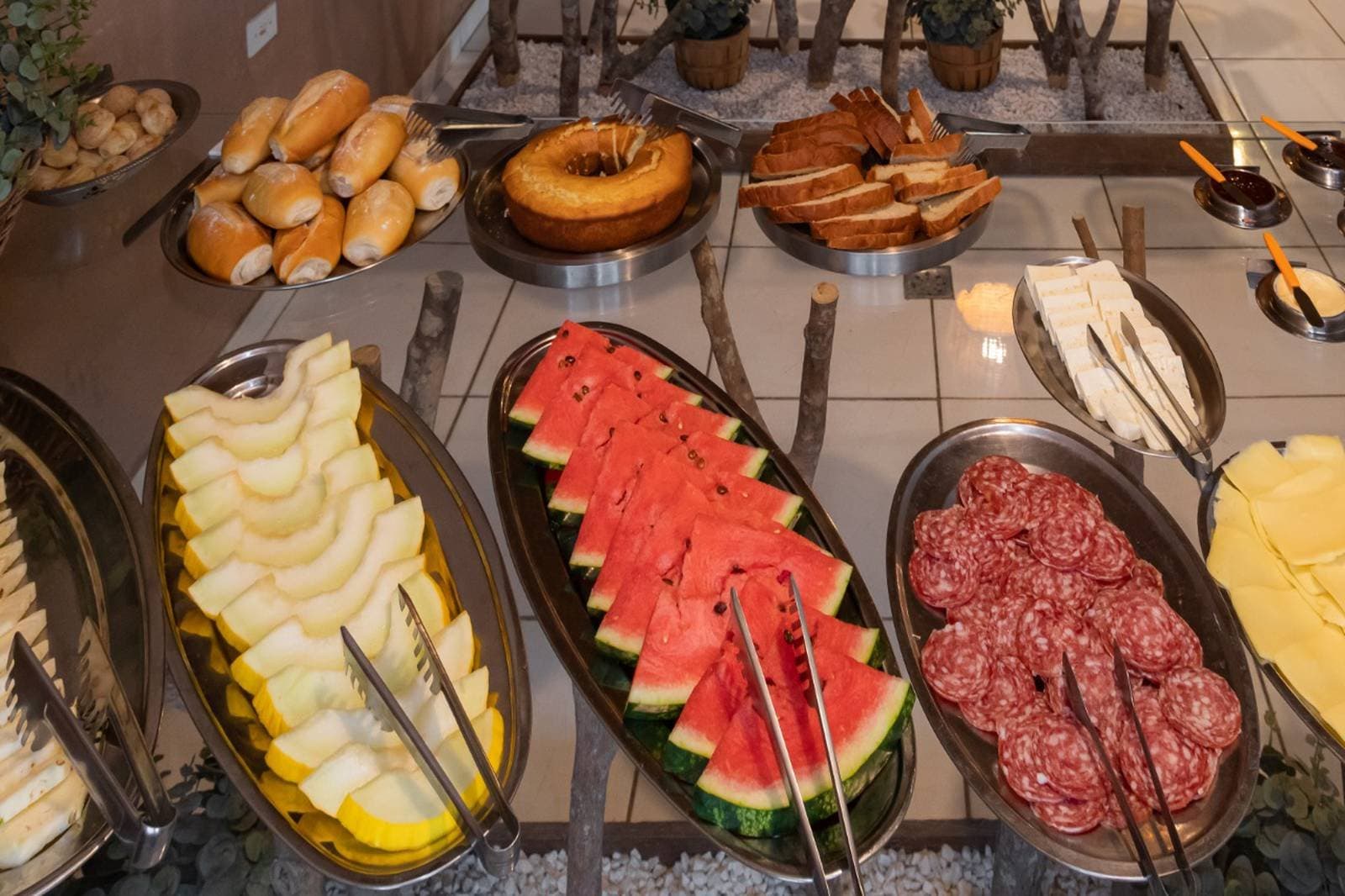 Wer früh aufbricht, braucht ein ordentliches Frühstück. Ein reichhaltiges Frühstücksbuffet auf einer Budget-Pantanal-Tour mit Wassermelonenscheiben, Honigmelone, Salamischeiben, Käse, Brötchen, einem Ringkuchen, Toast und Dipsaucen in Edelstahlschalen