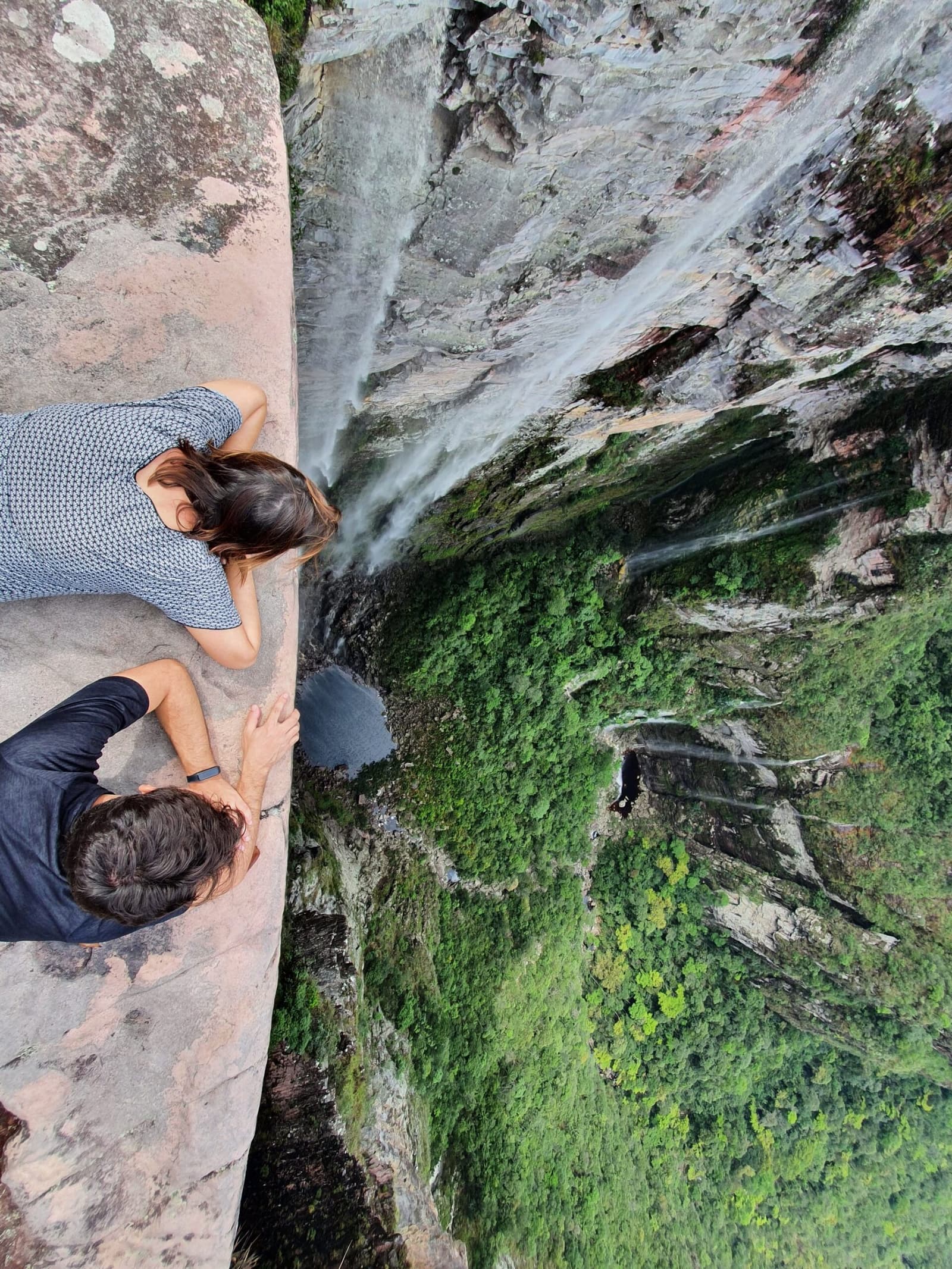 Bauch auf dem Stein, Blick nach unten, so sieht Cachoeirão aus nächster Nähe aus. Photo: Lucas Neves Zwei Trekker liegen flach auf einem Sandsteinfelsen am Rand der Chapada Diamantina und blicken senkrecht eine mit Wasserfällen durchzogene Felswand in eine grüne Schlucht hinab