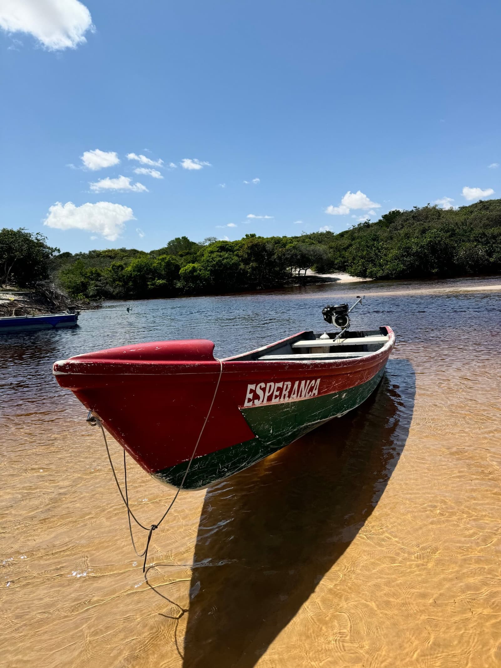 Esperança encalhada na beira do rio, pronta para levar você ao próximo horizonte. Um barco de madeira vermelho e verde chamado Esperança amarrado em uma margem de areia, com vegetação de mangue e céu azul ao fundo — parte do roteiro completo pelos Lençóis Maranhenses