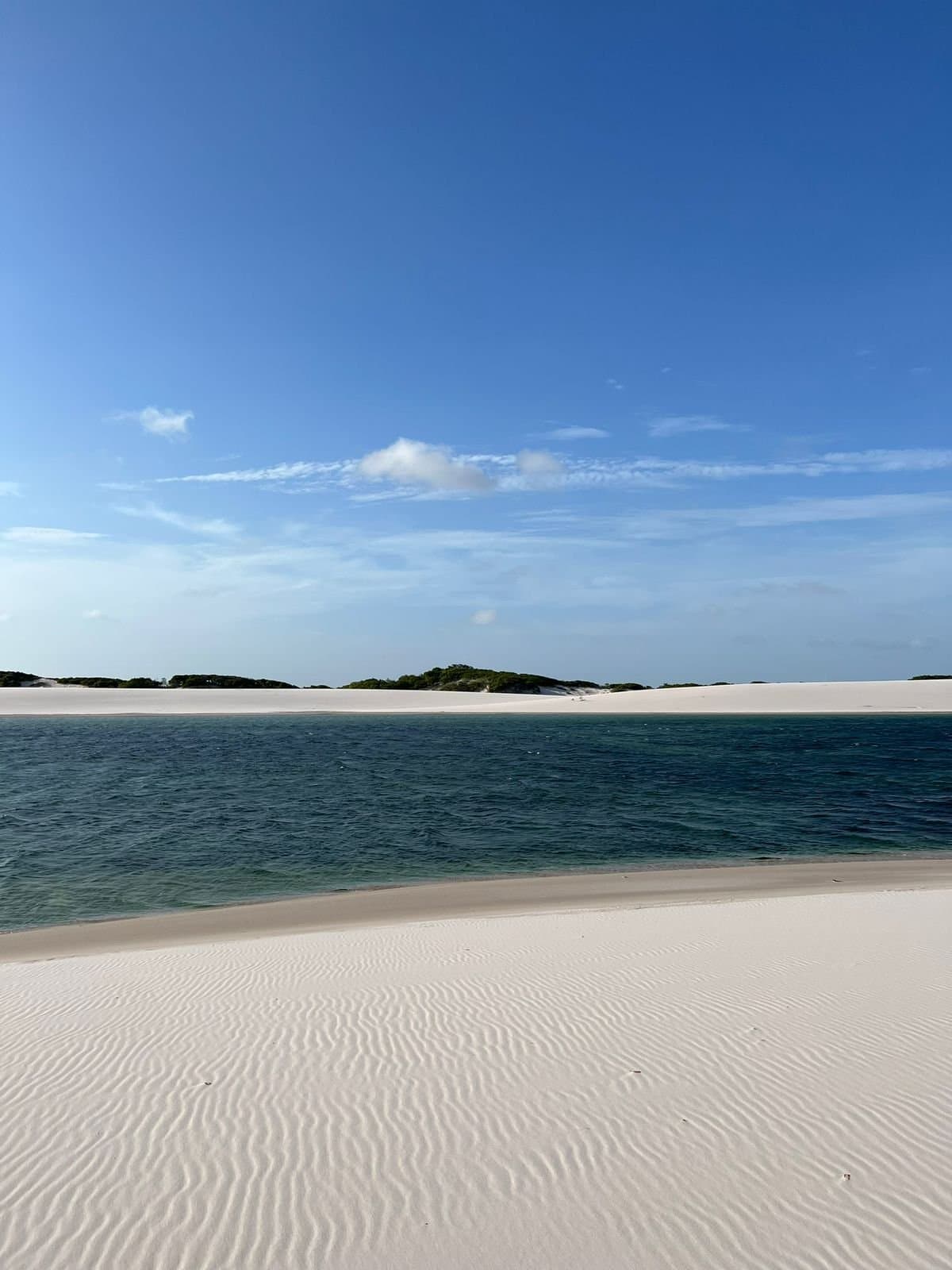 Le genre d'endroit qu'on hésite à montrer, de peur que tout le monde y coure. Large lagune d'eau douce bordée de dunes de sable blanc ondulées sous un ciel bleu dégagé à Lençóis Maranhenses Santo Amaro, avec une végétation verte clairsemée sur la crête de dune au loin