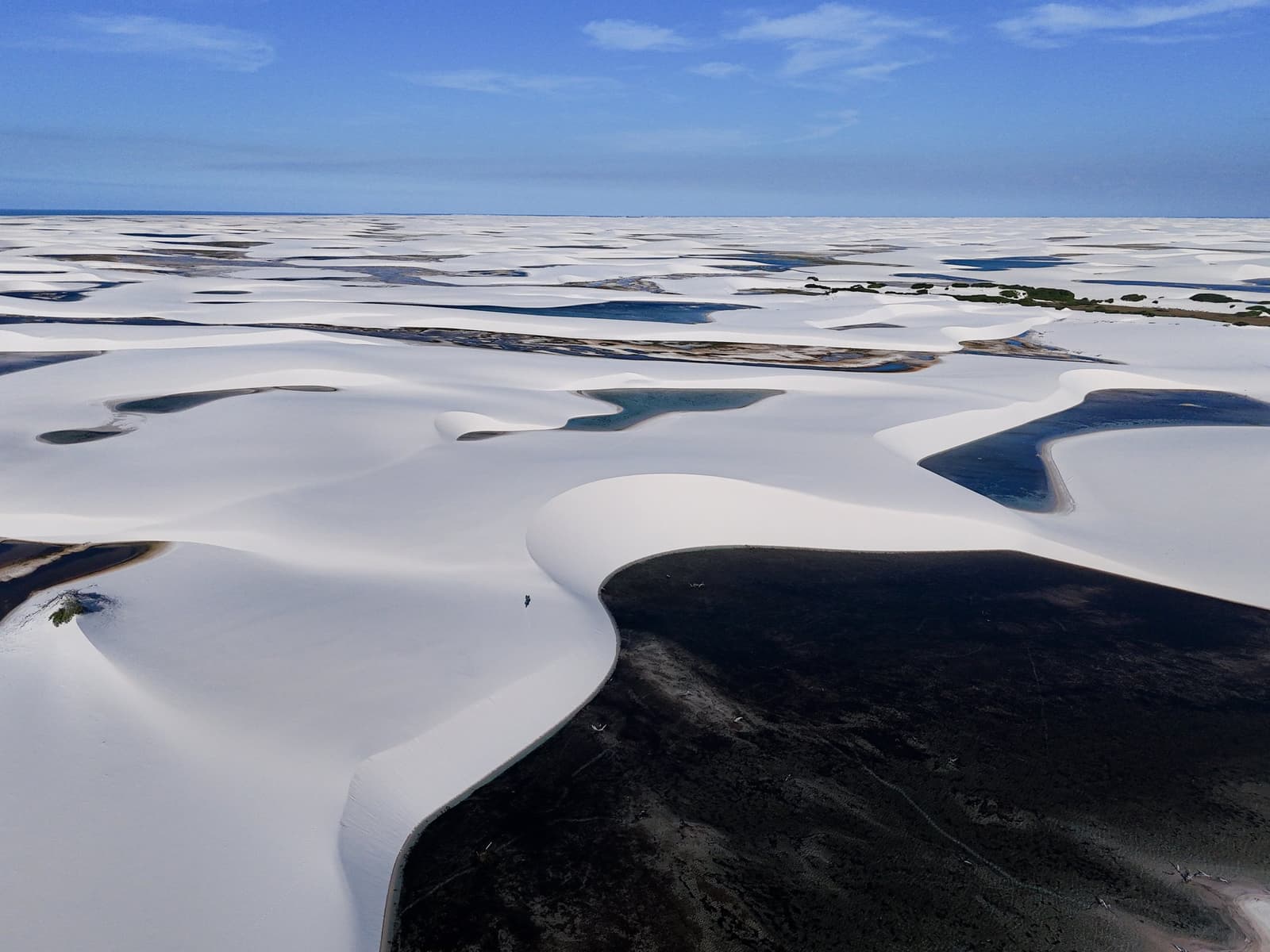 Uma pessoa, milhões de dunas. O parque tem um jeito próprio de colocar tudo em perspectiva. Vista aérea do trek pelos Lençóis Maranhenses saindo de Atins, mostrando imensas dunas de areia branca que se estendem até o horizonte, com lagoas de água doce escuras e turquesa entre as cristas das dunas sob um céu azul limpo, e uma figura humana diminuta visível sobre uma duna ao fundo