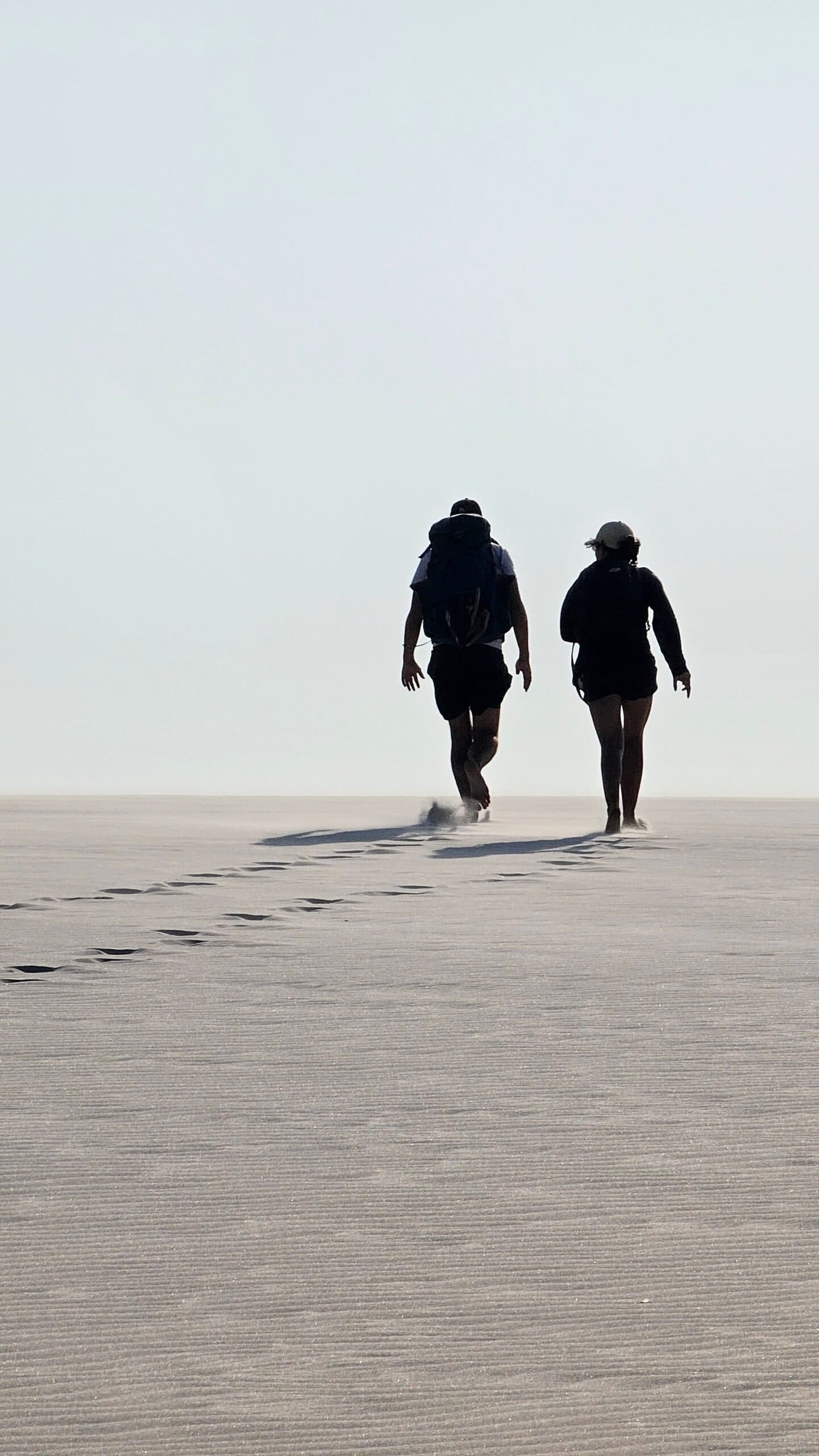 Areia branca até onde os olhos alcançam, e cada passo é só seu. Dois caminhantes vistos de costas atravessando uma vasta planície de areia branca durante o trek pelos Lençóis Maranhenses saindo de Atins, com suas pegadas se estendendo atrás deles