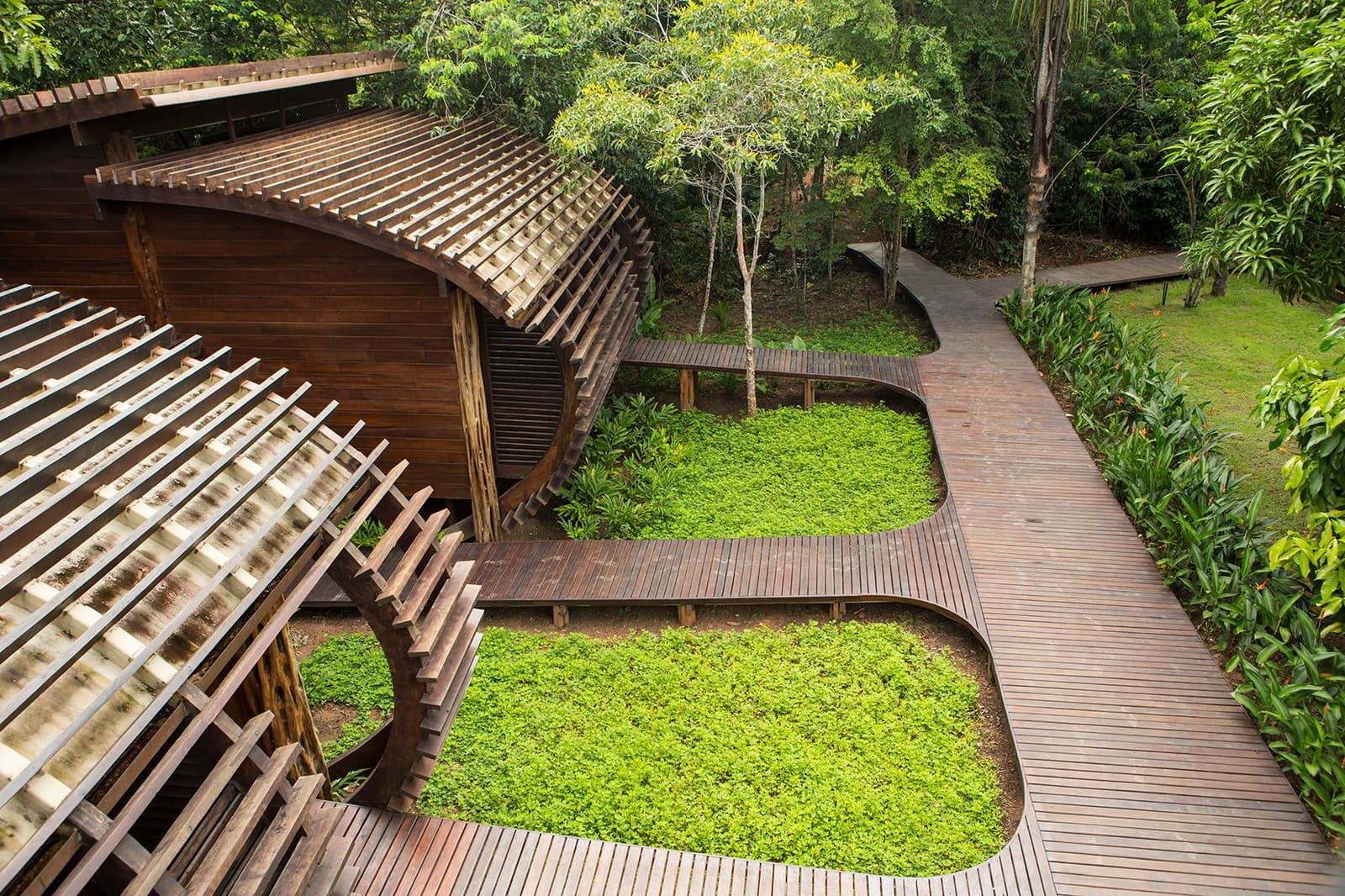 Quand l'architecture choisit de s'effacer devant la forêt. Vue en hauteur des promenades en bois dur sinueuses de Mirante do Gavião serpentant entre un tapis végétal vert luxuriant et un pavillon à lattes de bois, avec la canopée dense de la forêt tropicale en arrière-plan
