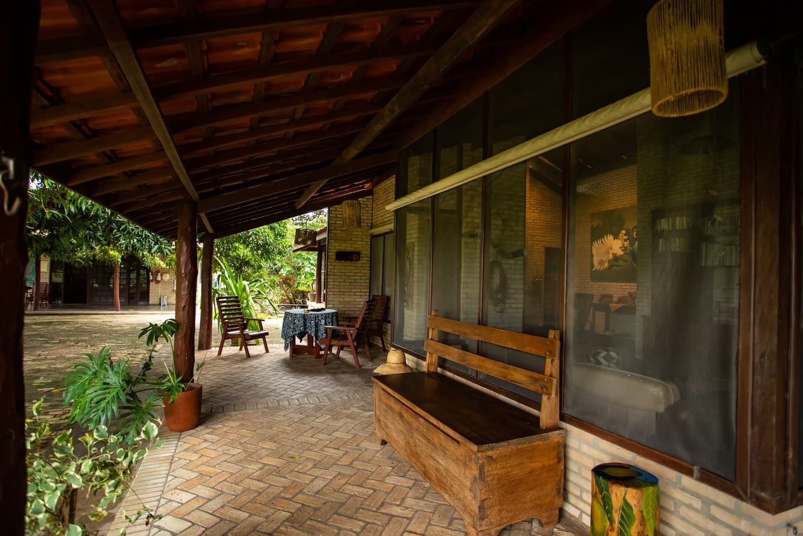 Posez vos affaires, le hamac peut attendre. La terrasse, elle, est prête. Véranda couverte en brique d'un lodge de safari dans le sud du Pantanal, avec un banc et un coffre en bois rustiques au premier plan, des chaises en bois autour d'une table et un jardin verdoyant en fond de cour