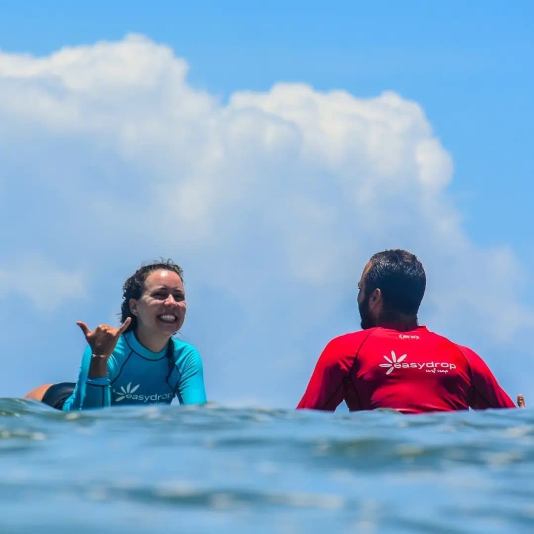 Die shaka zegt alles over hoe de les ging. Een glimlachende cursiste in een turquoise EasyDrop rashguard geeft haar instructeur een shaka-teken tijdens surflessen in Itacaré, terwijl ze allebei op hun boards zitten in de open oceaan onder een blauwe lucht.