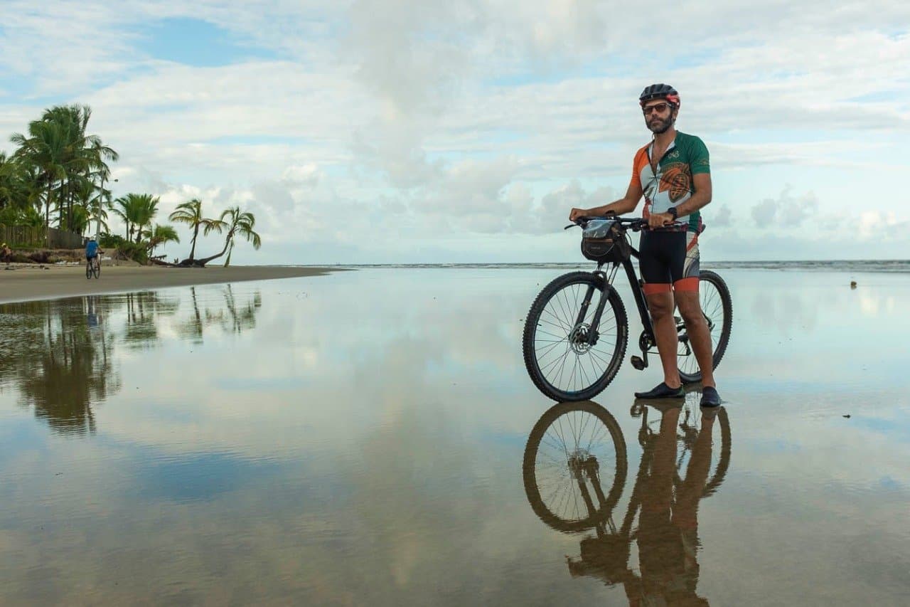 Areia molhada, céu espelhado, e a estrada toda ainda por vir. Um ciclista com camisa verde e laranja para com sua bicicleta de montanha em uma praia plana com lâmina d'água refletindo nuvens e palmeiras, com outro ciclista ao fundo — parte de um passeio de ciclismo na Bahia pela costa sul.